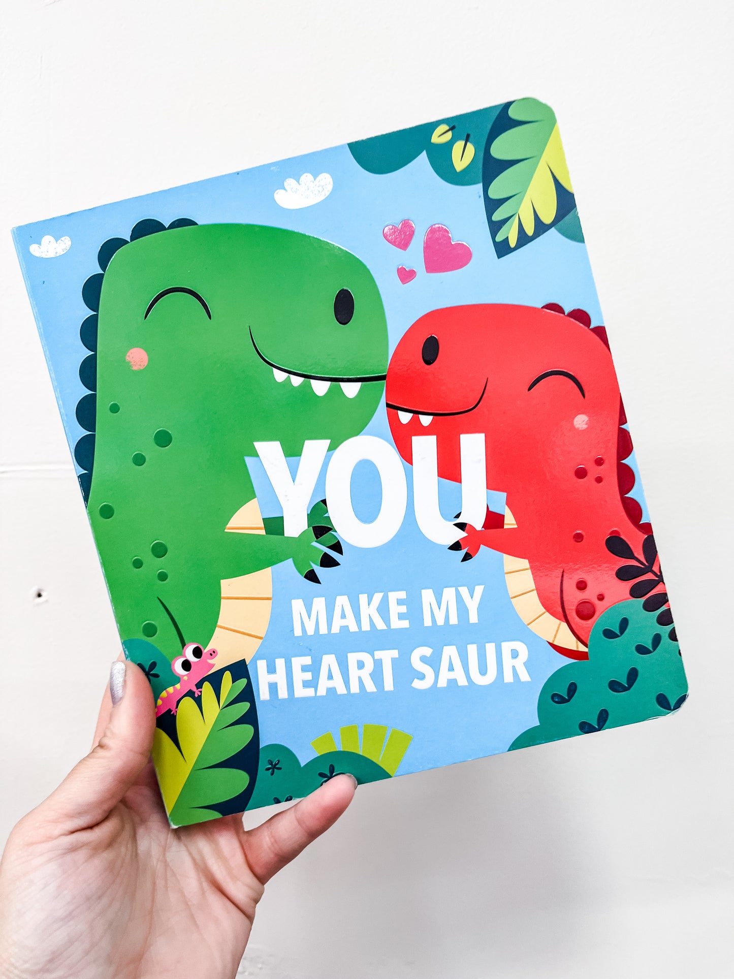 A hand holds the "You Make My Heart Saur Book" by Simon & Schuster, featuring two smiling cartoon dinosaurs facing each other with leaves and hearts—a sweet toddler board book perfect for dinosaur fans.