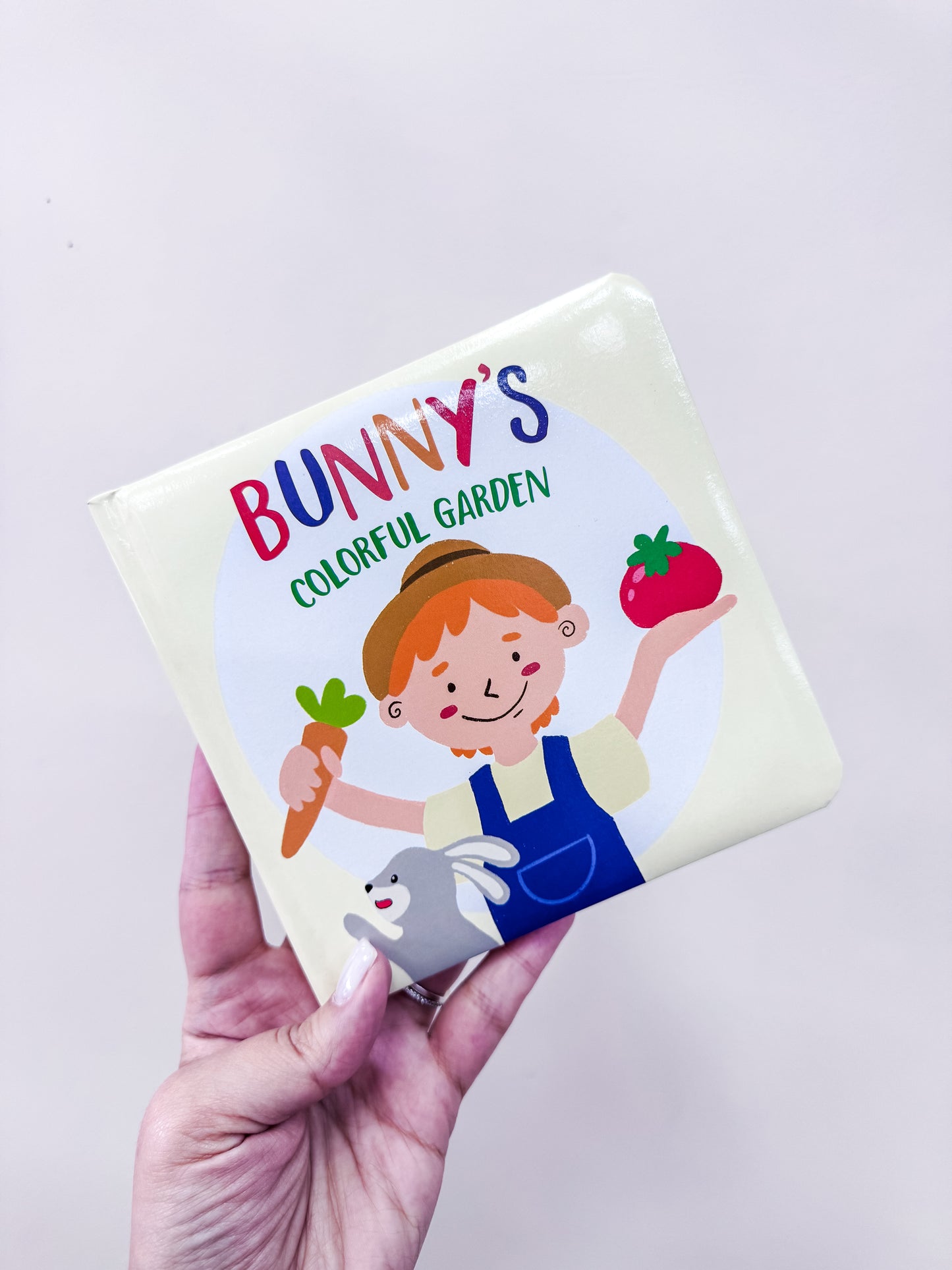 A hand holds the "Colorful Vegetable Garden Board Book" by Tiny Tot Bundles, featuring a child with a carrot and strawberry, and a smiling bunny at the bottom.