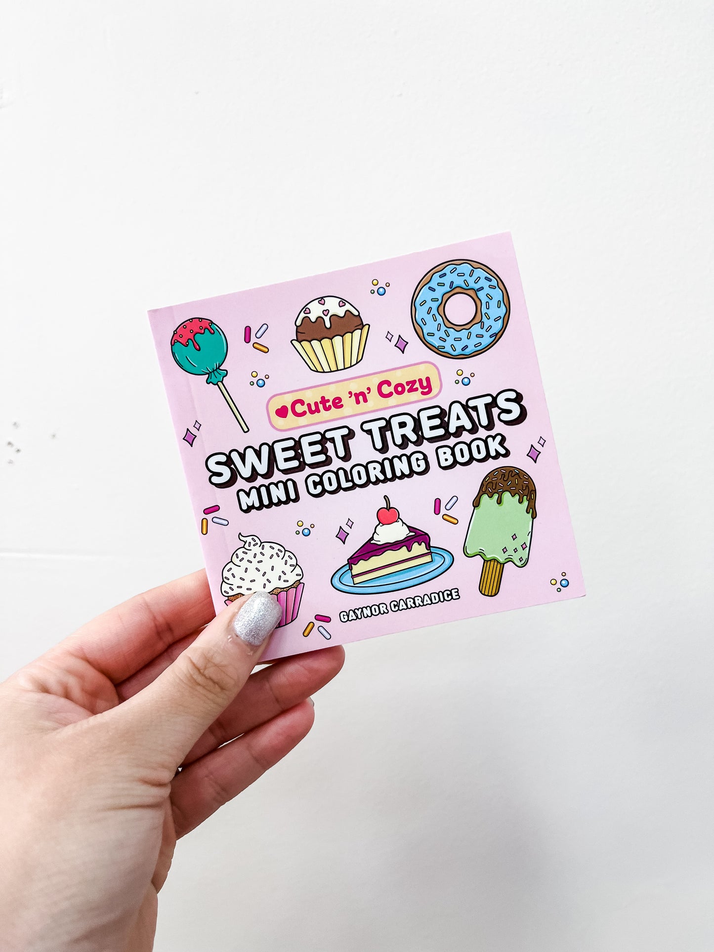 A hand with silver nail polish holds the Simon & Schuster Sweet Treats Mini Coloring Book—a portable book featuring cupcakes, donuts, cake, and ice cream on the cover.