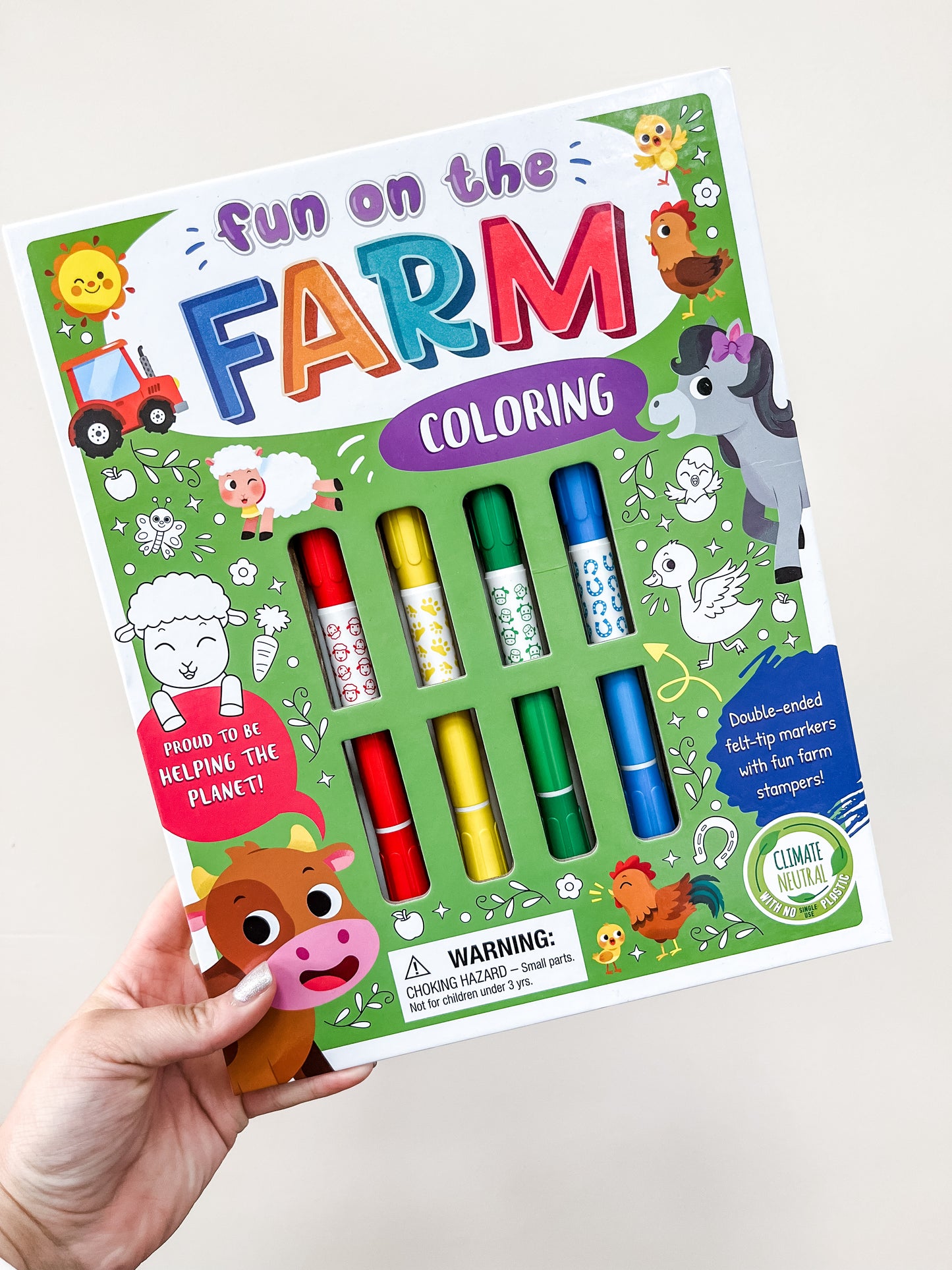 A hand holds the Simon & Schuster Fun On the Farm Coloring Set Book, which includes six colorful markers and farm animal illustrations like a dog, chicken, and cow on a playful green background.