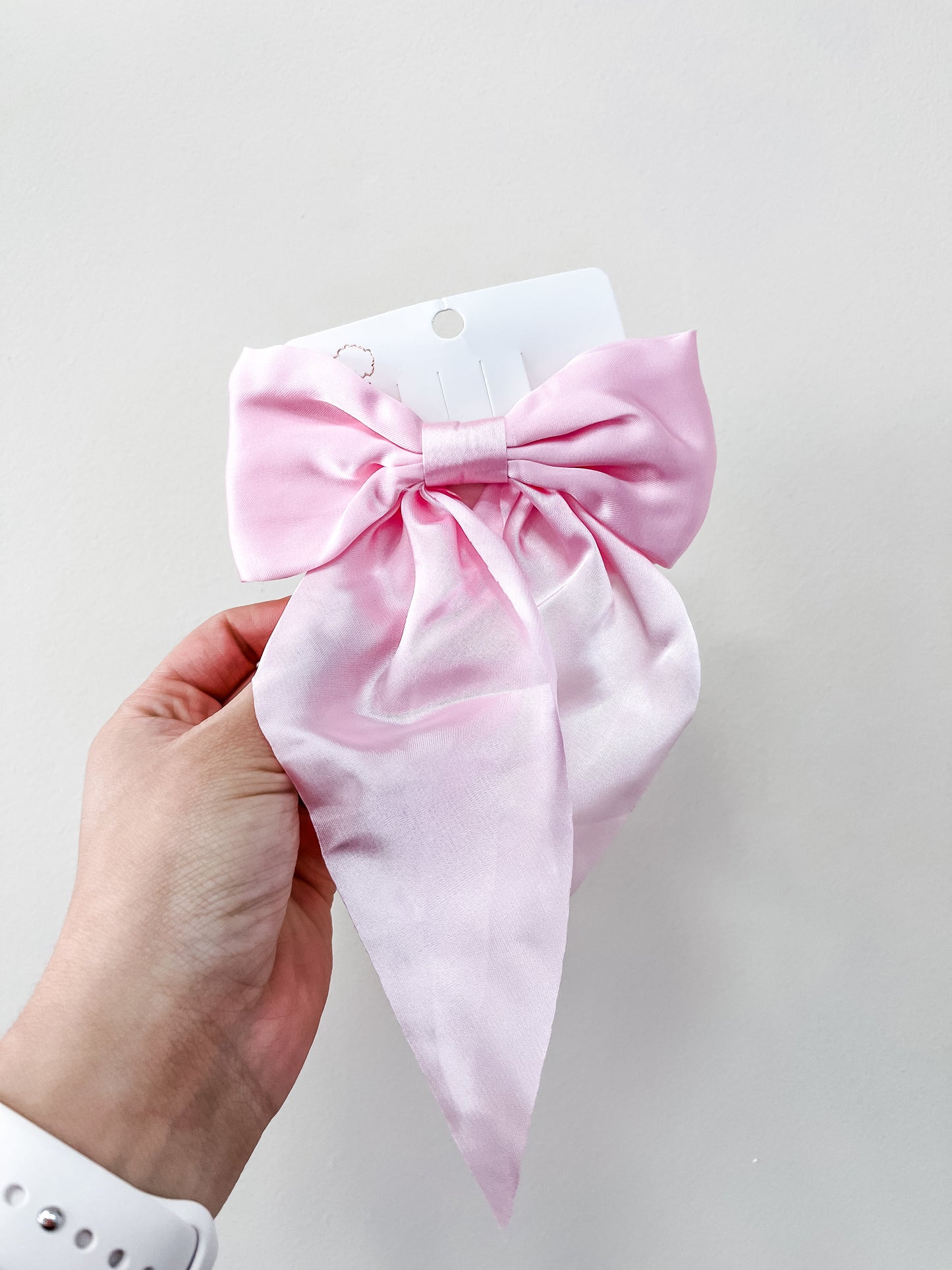 A hand holds the Sweetheart Statement Hair Clip by love & repeat—a large, light pink satin bow with long, flowing tails and a shiny finish—displayed on a white card against a plain light background.