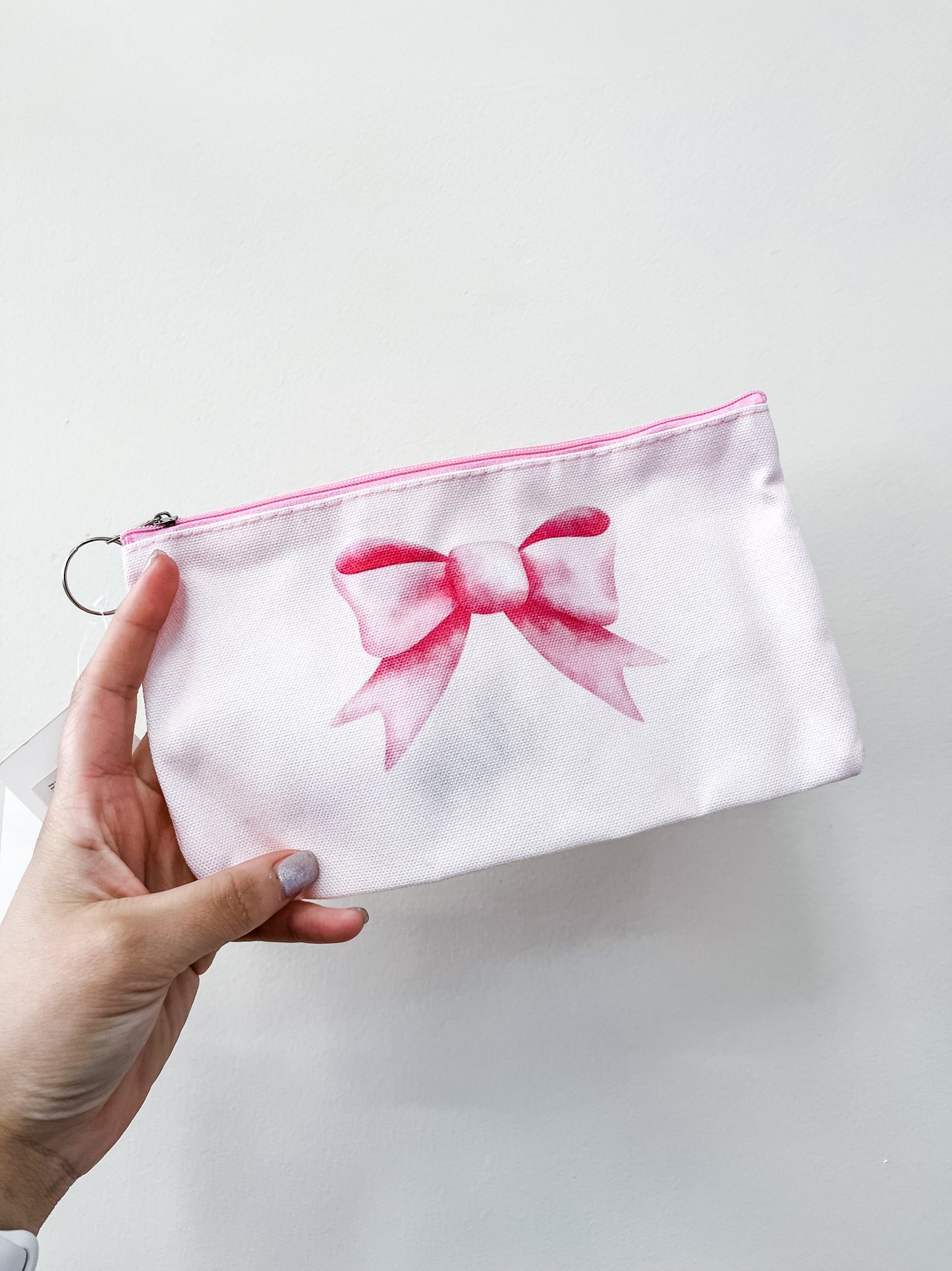 A hand holds the love & repeat Pretty in Bows Zipper Pouch, a light pink pouch with a silver keyring and a large pink watercolor-style bow on the front, set against a plain white background.
