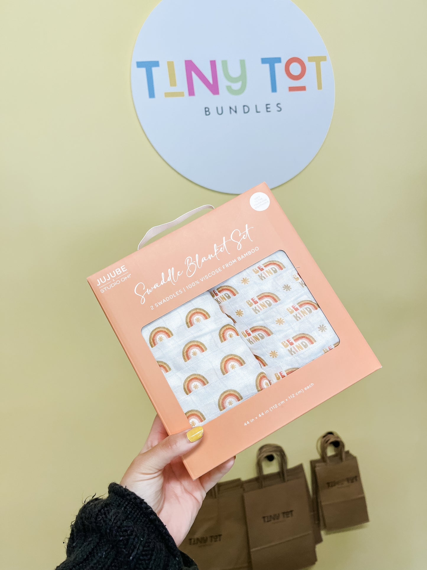 A hand holds a Tiny Tot Bundles Swaddle Blanket Set - Be Kind Rainbows in front of a wall sign and paper shopping bags, both featuring the Tiny Tot Bundles logo.