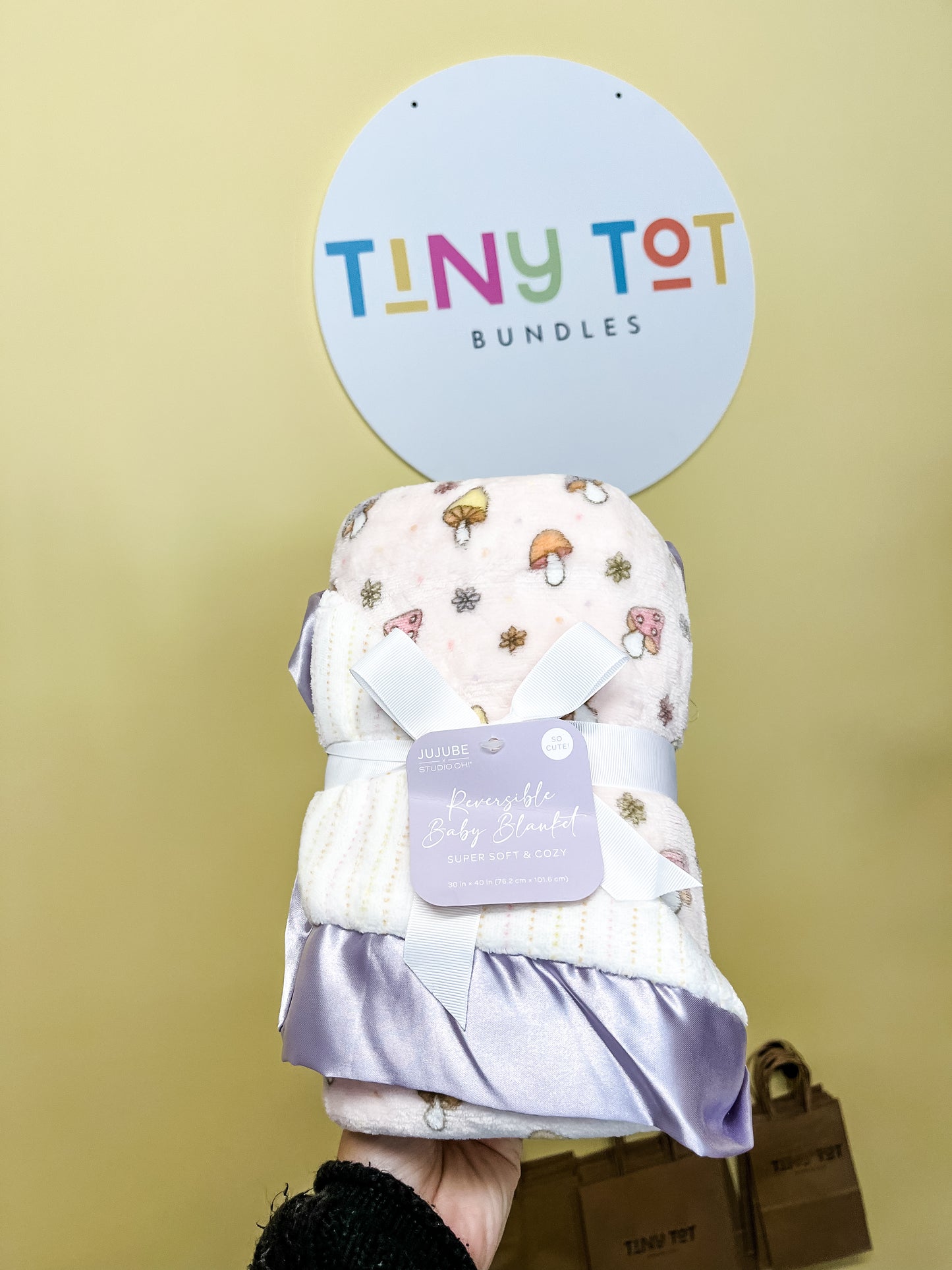 A hand holds the Reversible Baby Blanket - Mushy Love from Tiny Tot Bundles, featuring a soft, light purple mushroom and floral print, tied with a ribbon. A circular Tiny Tot Bundles sign is visible in the yellow background.