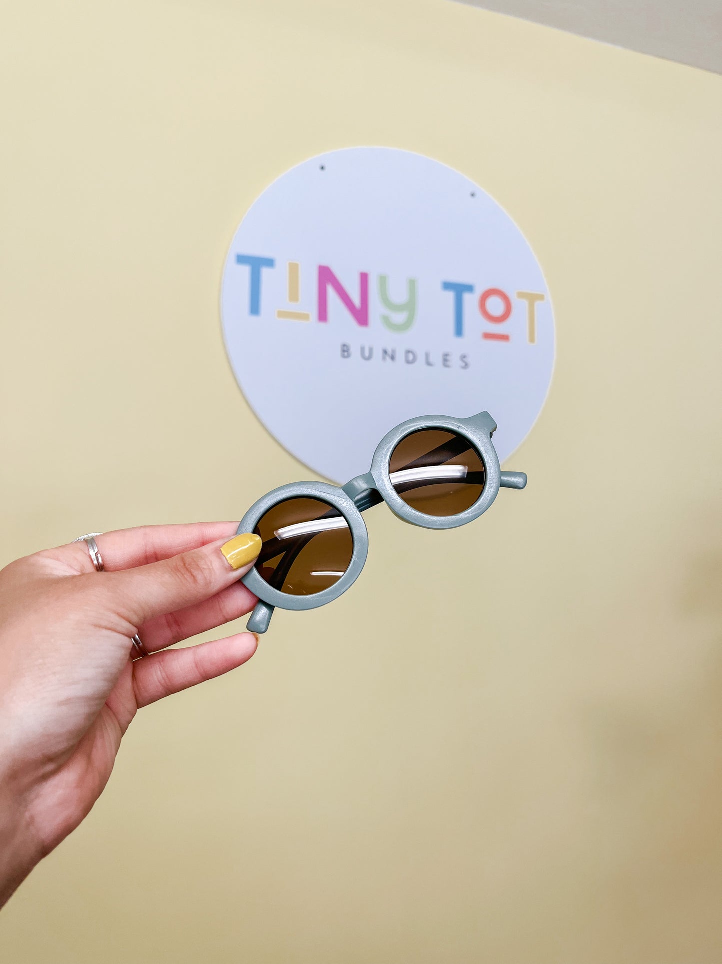 A hand with yellow nail polish holds up Modern Sunglasses by Tiny Tot Bundles in front of a colorful sign displaying the brand’s name.