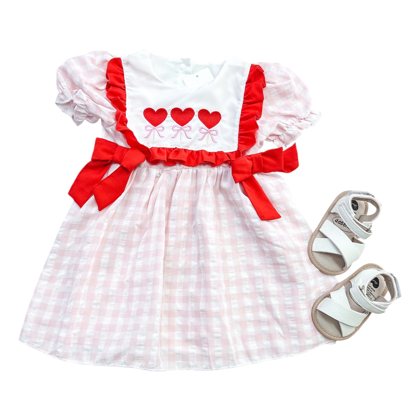 The Sweetheart Trio Dress by Tiny Tot Bundles features pink and white checks, red bows, and three red hearts on the chest, paired perfectly with white toddler sandals.