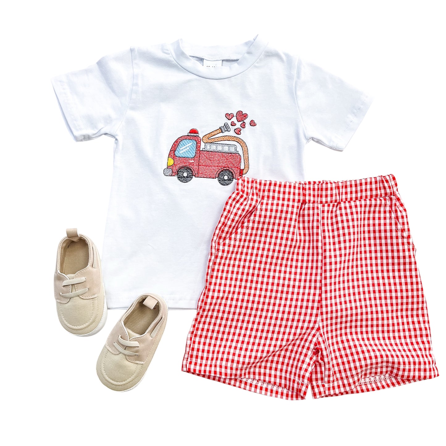The Tiny Tot Bundles Heart Hero Firetruck Set includes a white T-shirt with a red fire truck and hearts, red and white checkered shorts, and beige soft-soled shoes, displayed on a white background.