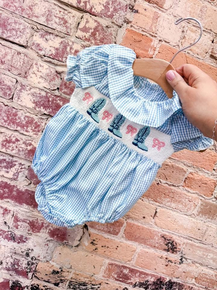 A hand holds the Tiny Tot Bundles Blue Smocked Cowgirl Romper with puff sleeves, a ruffled collar, and boot and pink bow embroidery, set against a rustic brick wall.