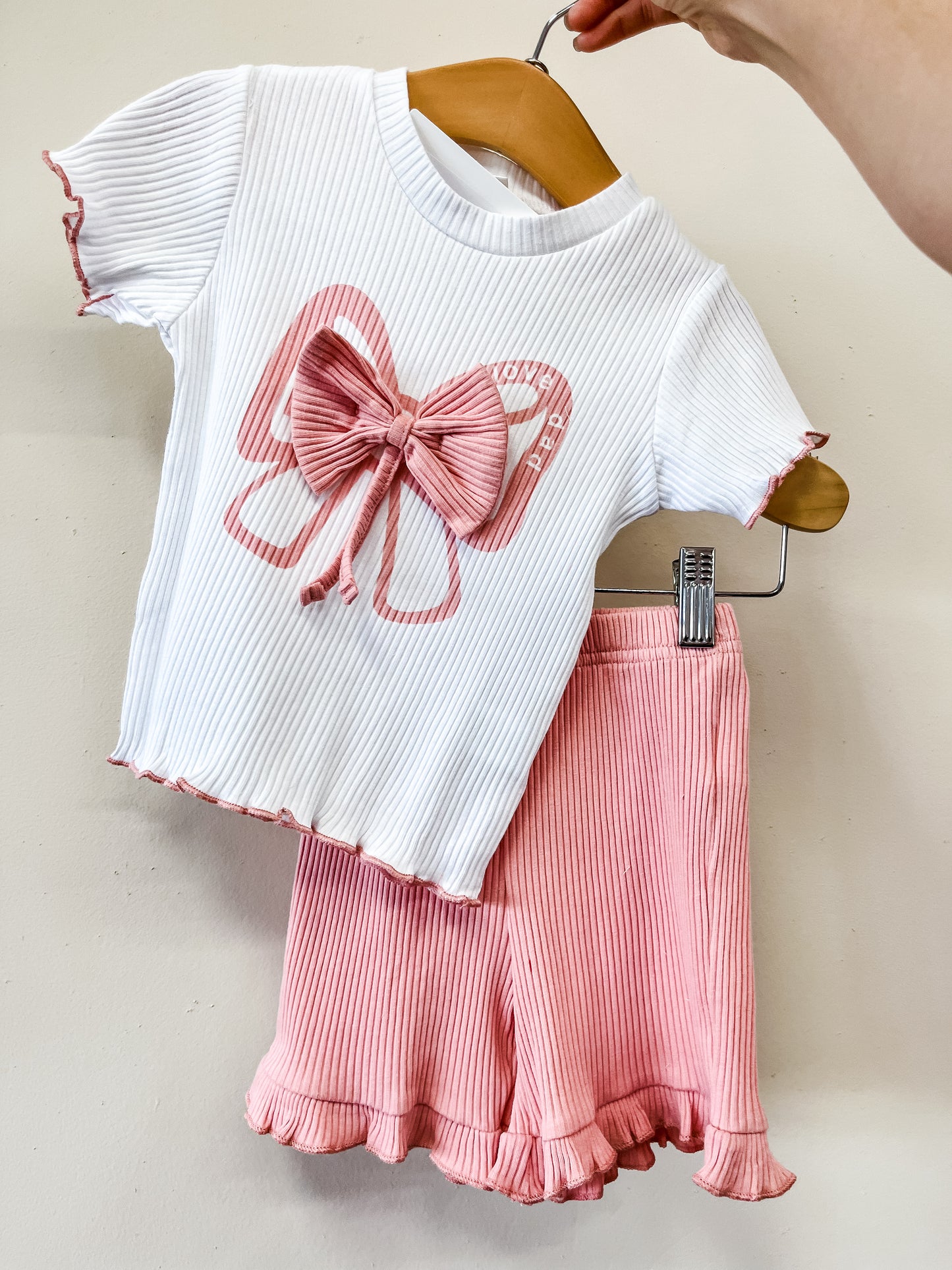 A hand displays Tiny Tot Bundles' Lovely Bow Ribbed Set: a white ribbed short-sleeve top with a pink bow and butterfly design, paired with pink ribbed shorts with ruffled hems. The background is plain off-white.