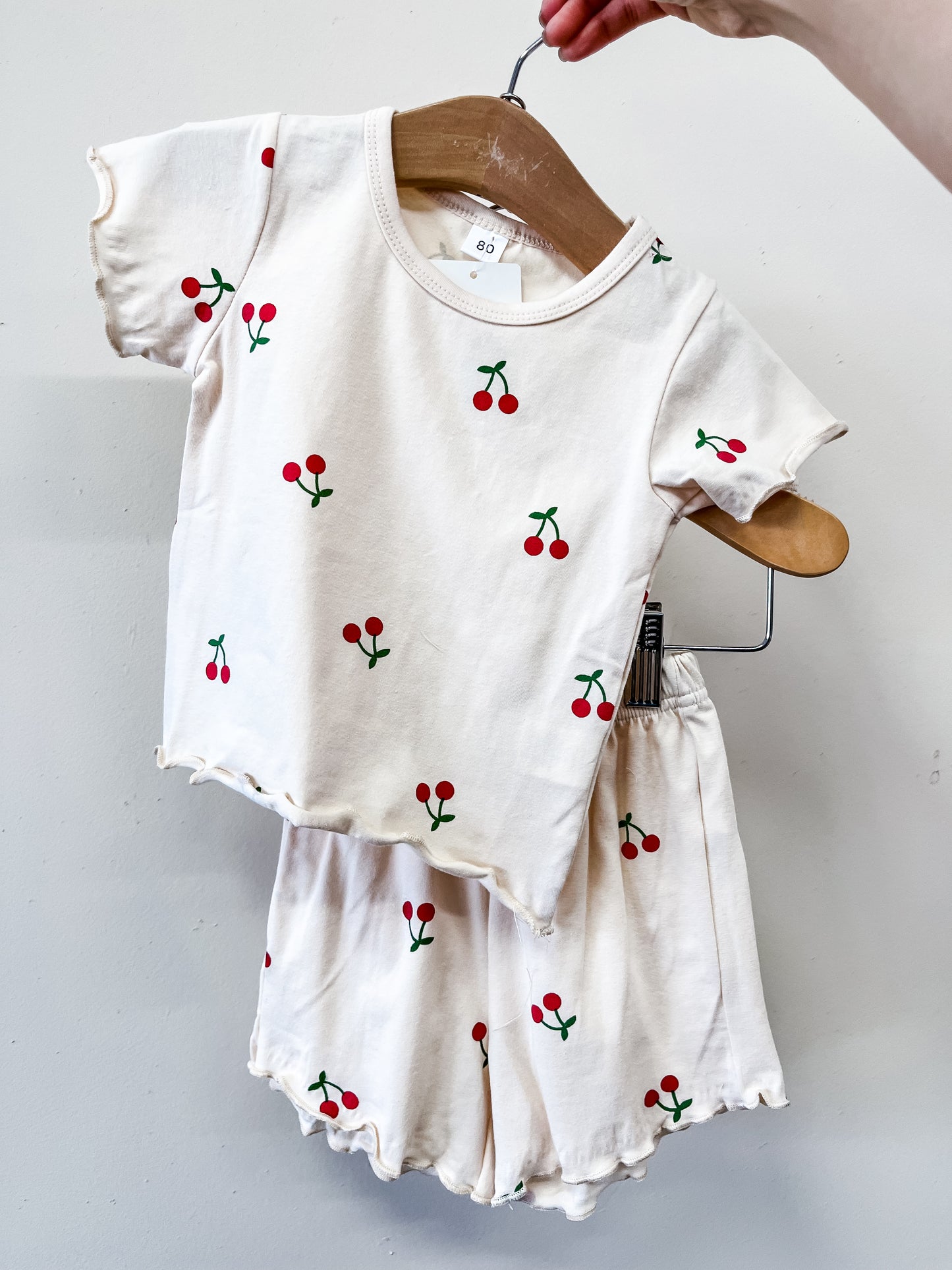The Cherry Picked Lounge Set by Tiny Tot Bundles features a cream-colored toddler short-sleeve shirt and matching cherry print shorts, displayed on a wooden hanger against a white background.