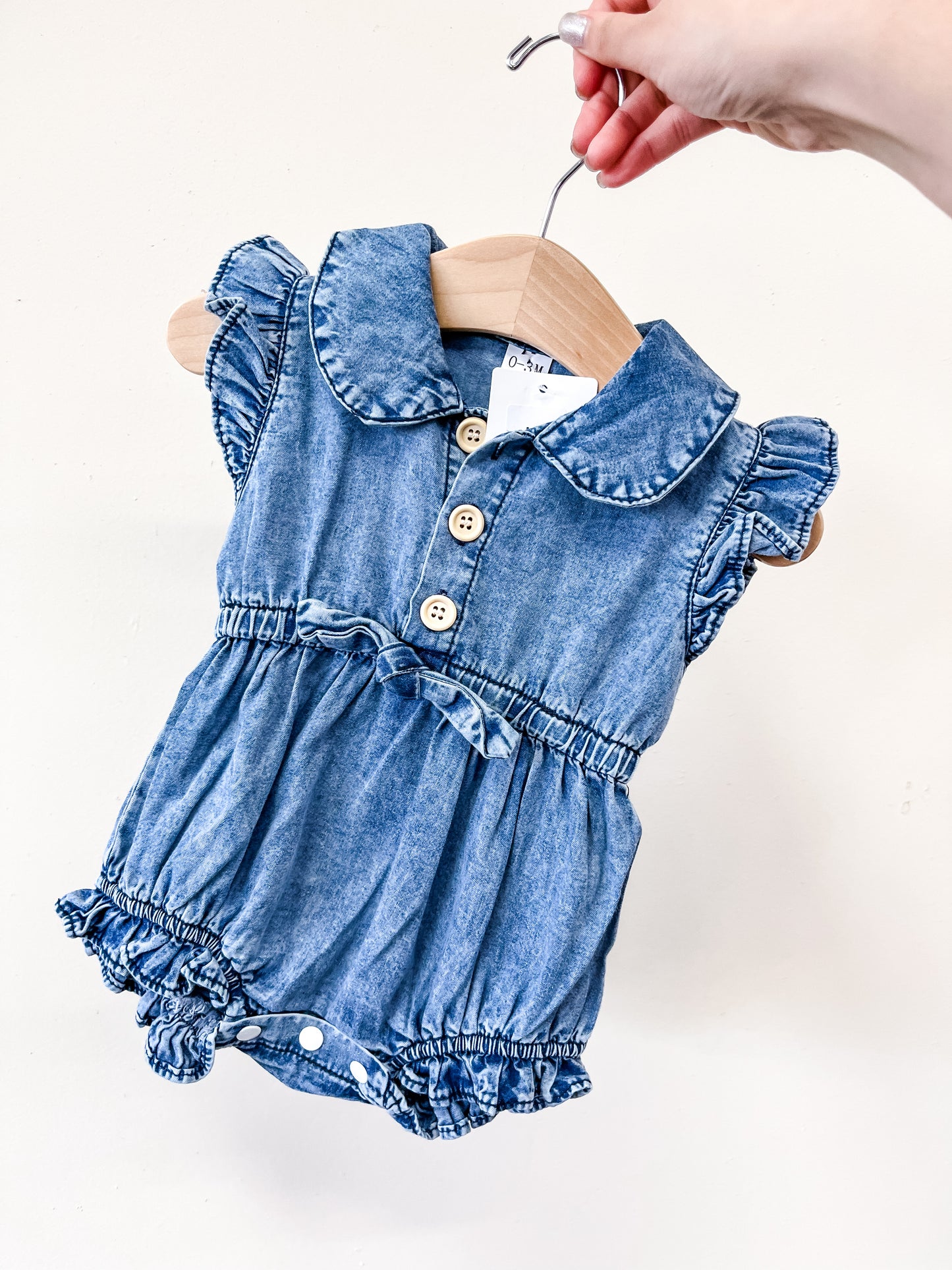 A hand holds a hanger with the Tiny Tot Bundles Denim Ruffle Romper—featuring blue denim, ruffled sleeves, a Peter Pan collar, wooden buttons, waist bow, and elasticized leg openings.