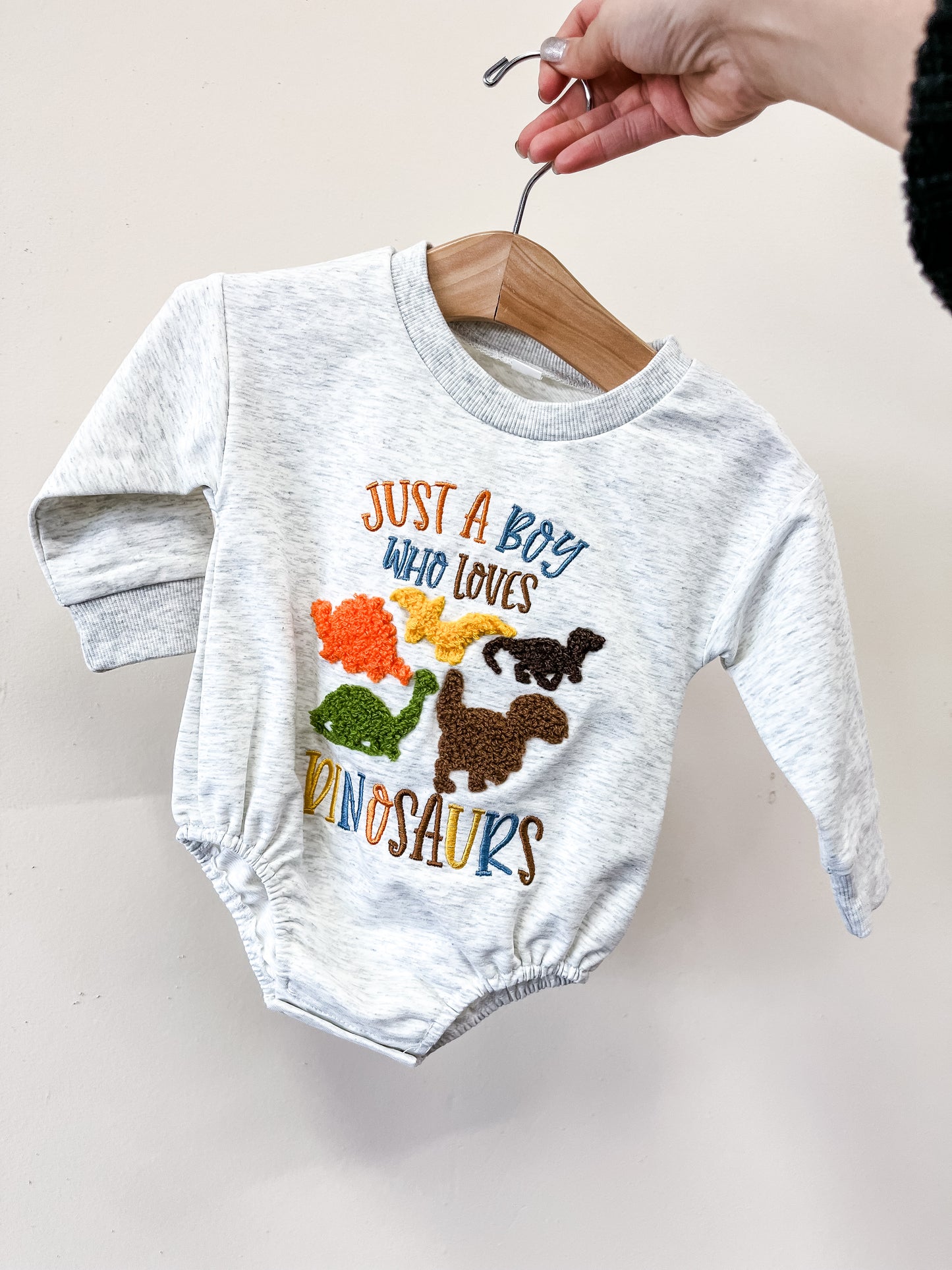 A hand holds a hanger with the Tiny Tot Bundles Dino Lover Bubble, a light gray baby onesie featuring colorful dinosaur graphics and "Just a boy who loves dinosaurs" embroidered on the front.