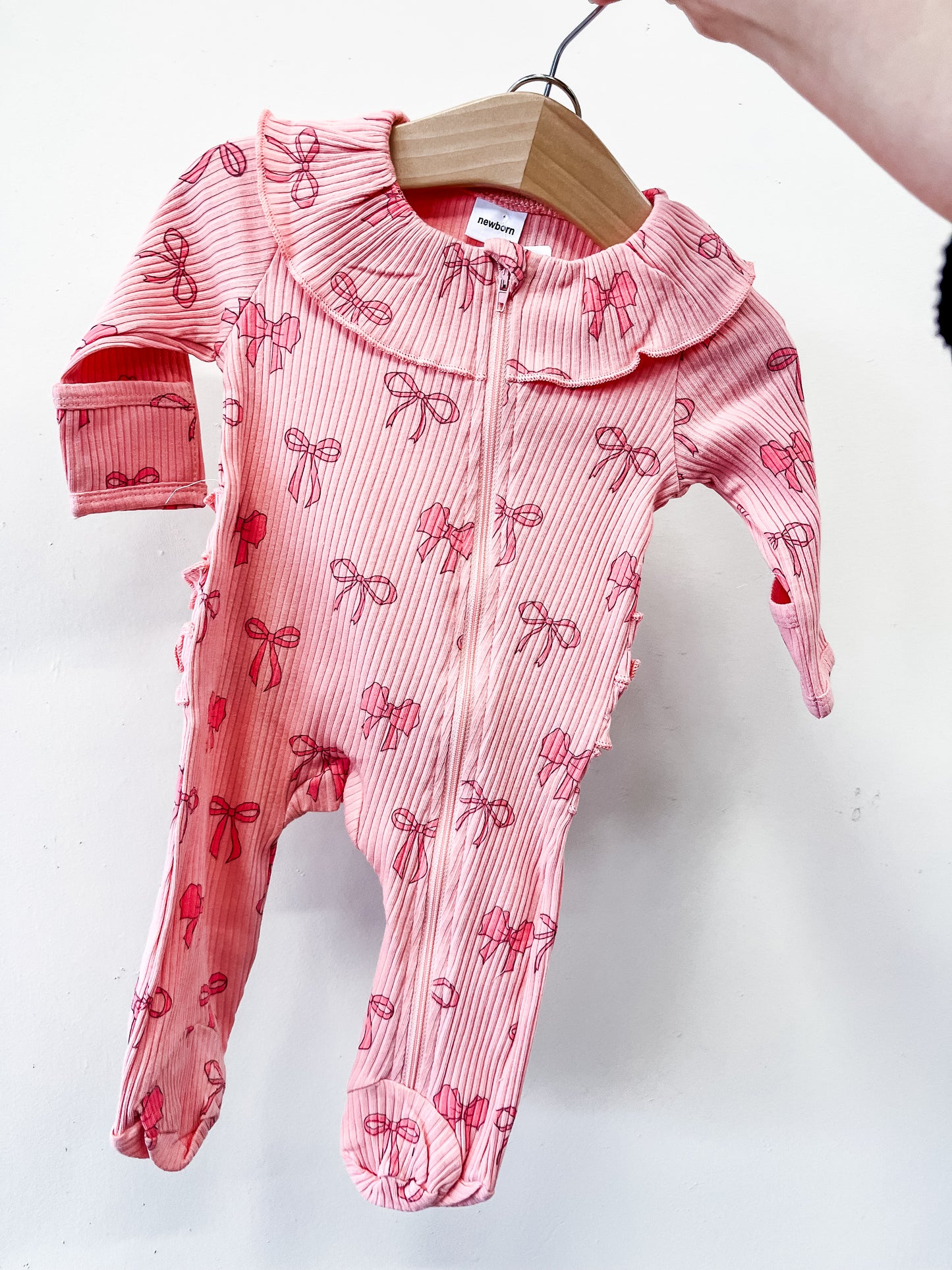 A hand holds the Bow Darling Ruffle Footie by Tiny Tot Bundles, a pink, long-sleeve onesie with ruffled collar, zipper front, and all-over bow print, displayed on a wooden hanger against a white background.