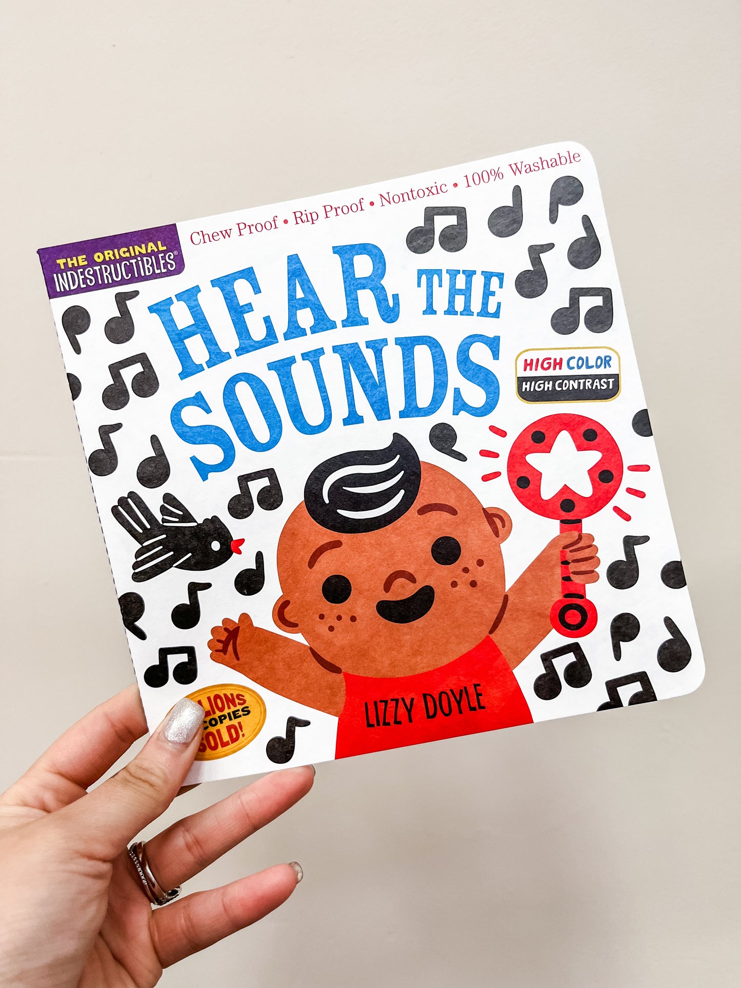 A hand holds Hear The Sounds Book by Hachette Book Group, featuring a smiling child with a rattle, musical notes, and a black bird on the cover. The book is chew proof and nontoxic.