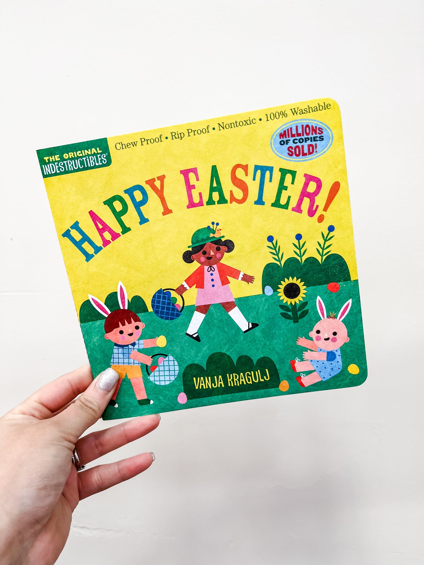 A hand holds the Happy Easter Book by Hachette Book Group. The cover features three cartoon characters—two bunnies and a girl—amid flowers, eggs, and bright spring colors.
