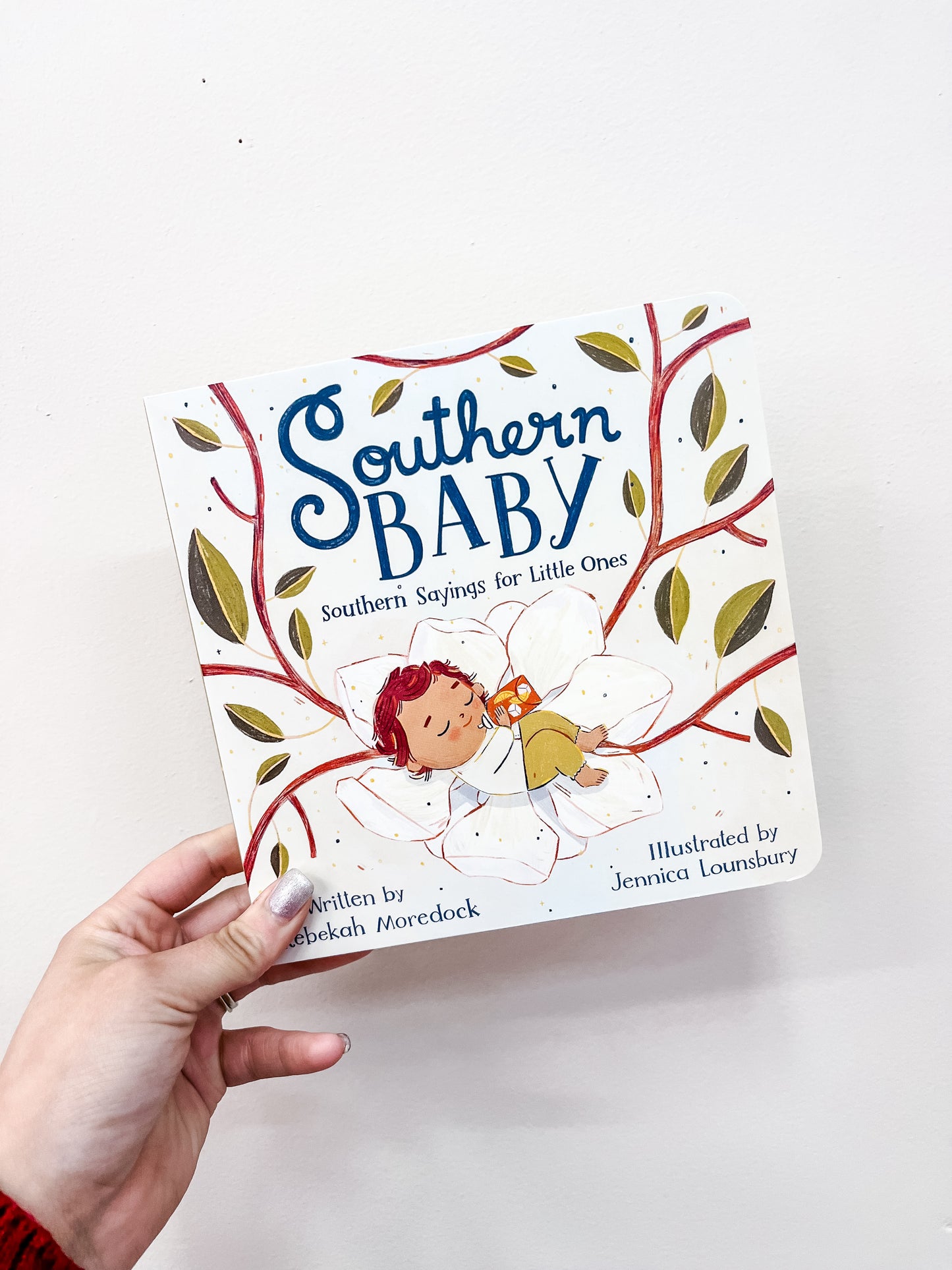 A hand holds the Southern Baby Book by Hachette Book Group, featuring an illustration of a baby sleeping on a white flower with branches and leaves in the background.