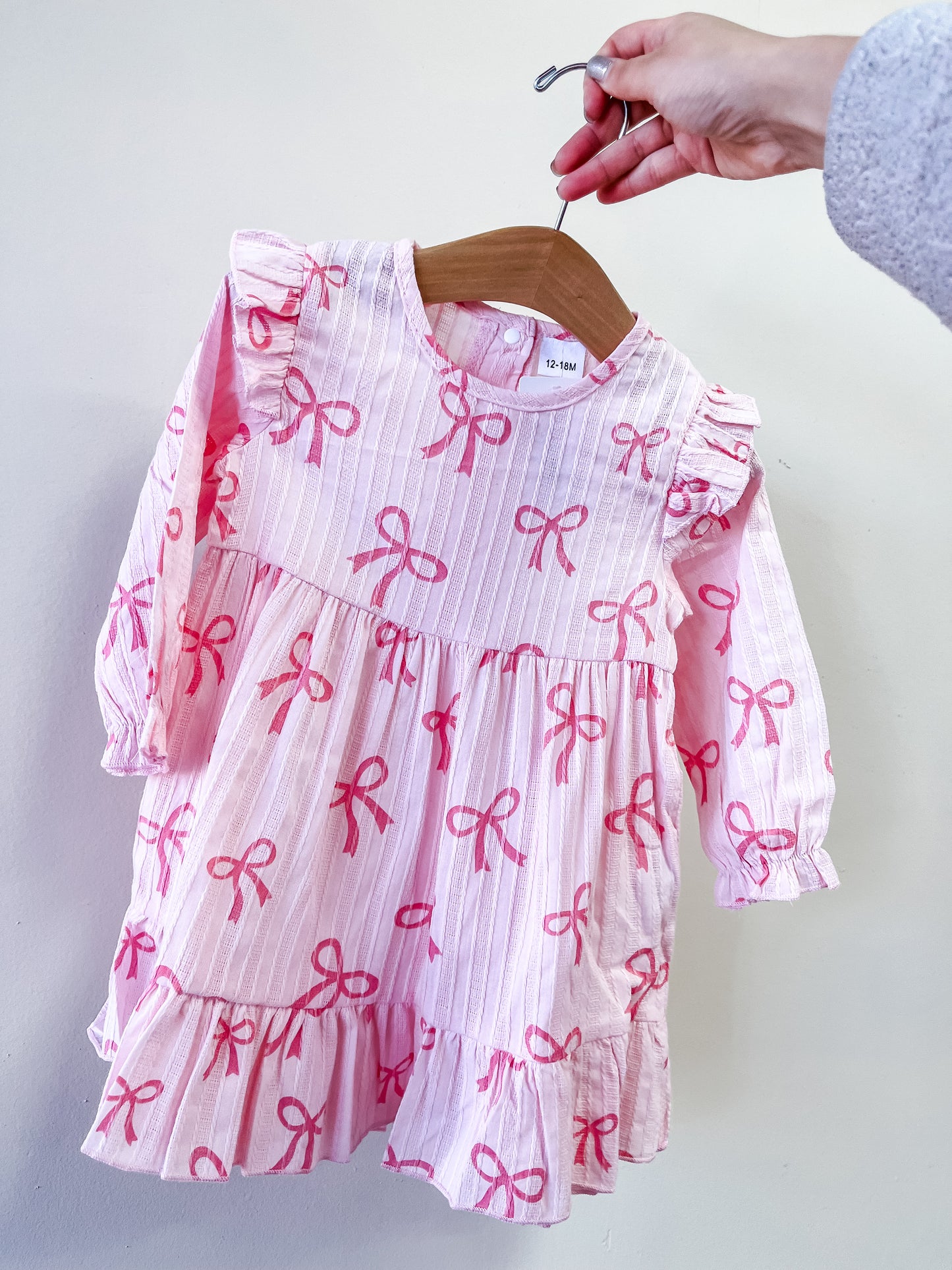 A hand displays the Tiny Tot Bundles Bow Bliss Dress, a pink long-sleeved baby dress with ruffles and darker pink bow patterns, shown on a wooden hanger against a plain light background.