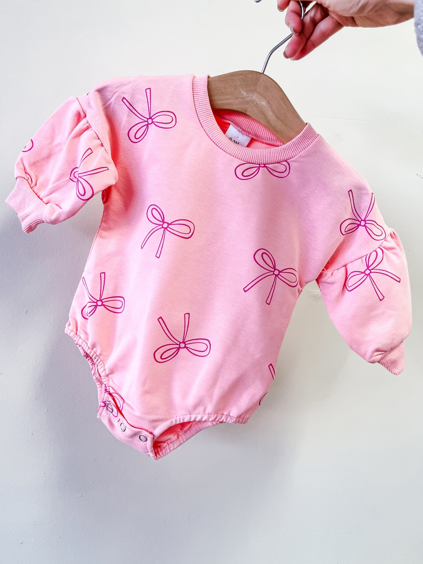 The Blush Bow Bubble by Tiny Tot Bundles, a pink long-sleeve baby onesie featuring simple outlined bows, is displayed on a wooden hanger held by hand.