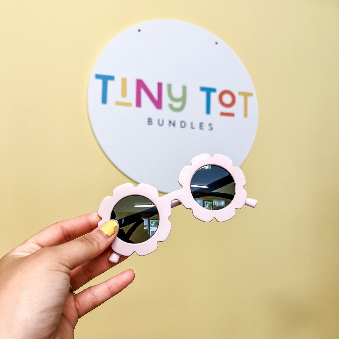 A hand with yellow nail polish holds Floral Sunglasses by Tiny Tot Bundles in front of a round sign that says Tiny Tot Bundles on a light yellow wall.