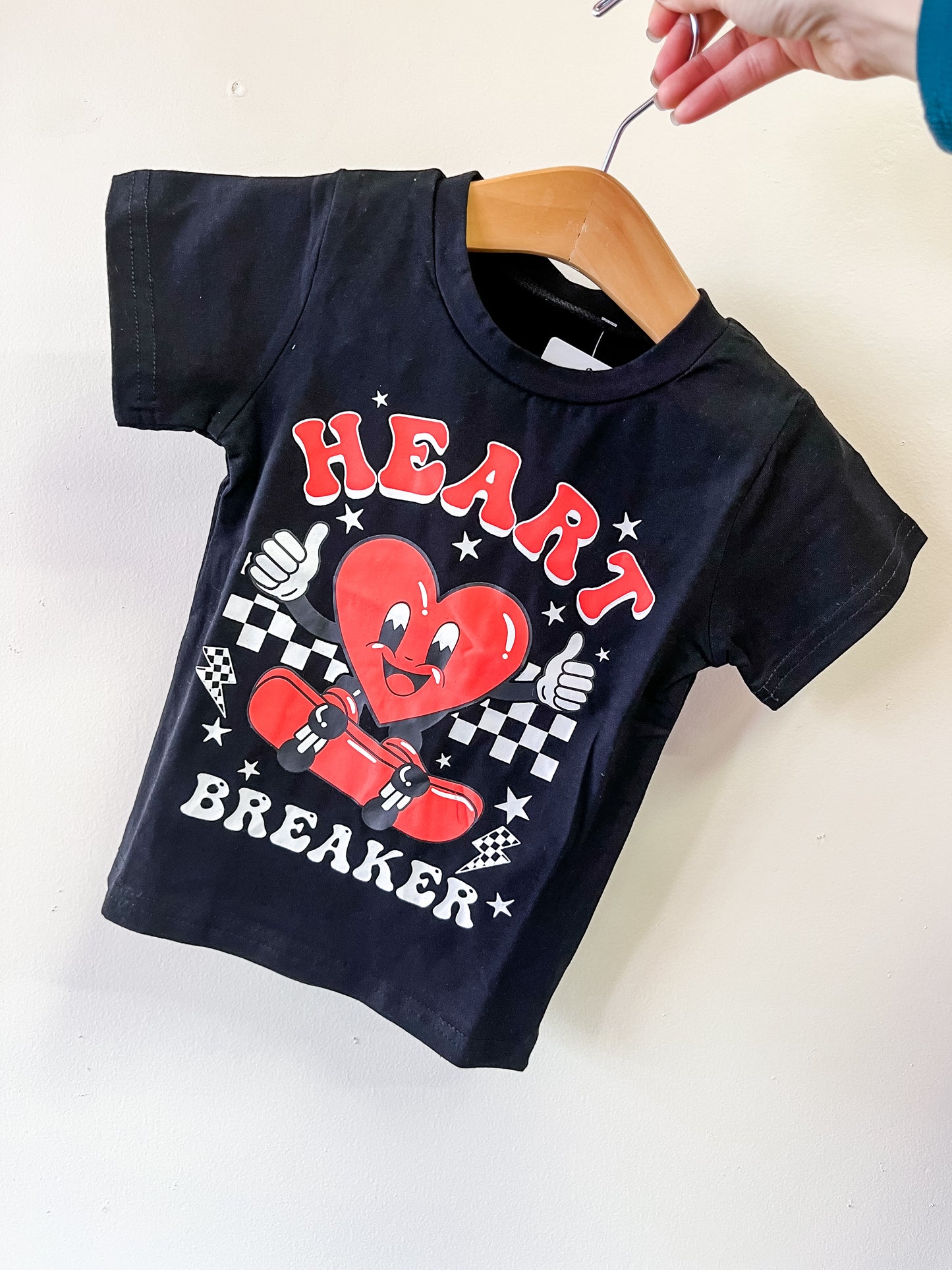 The Heart Breaker Graphic Tee by Tiny Tot Bundles is a black toddler t-shirt on a hanger, featuring a smiling cartoon heart on a skateboard, HEART BREAKER text, and a checkered flag design.