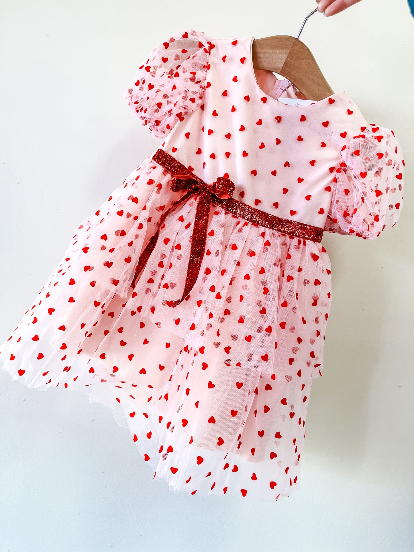 The Valentine's Tulle Dress by Tiny Tot Bundles is a pink toddler dress with red heart patterns, short puffy sleeves, and a red glittery waist ribbon, shown on a wooden hanger against a white background.