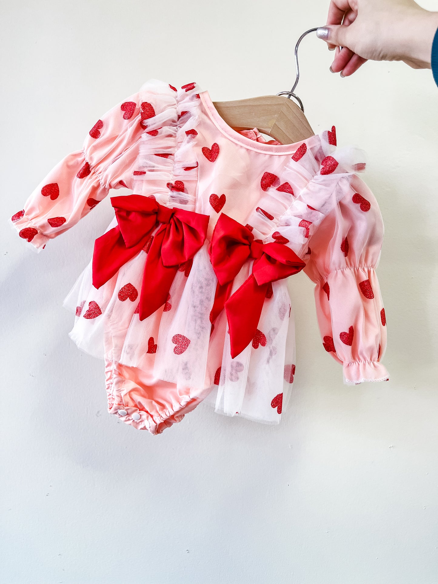 The Sweetheart Tulle Romper by Tiny Tot Bundles features pink fabric with red hearts, sheer ruffles, and two large red bows on the front. Displayed on a wooden hanger against a white background.