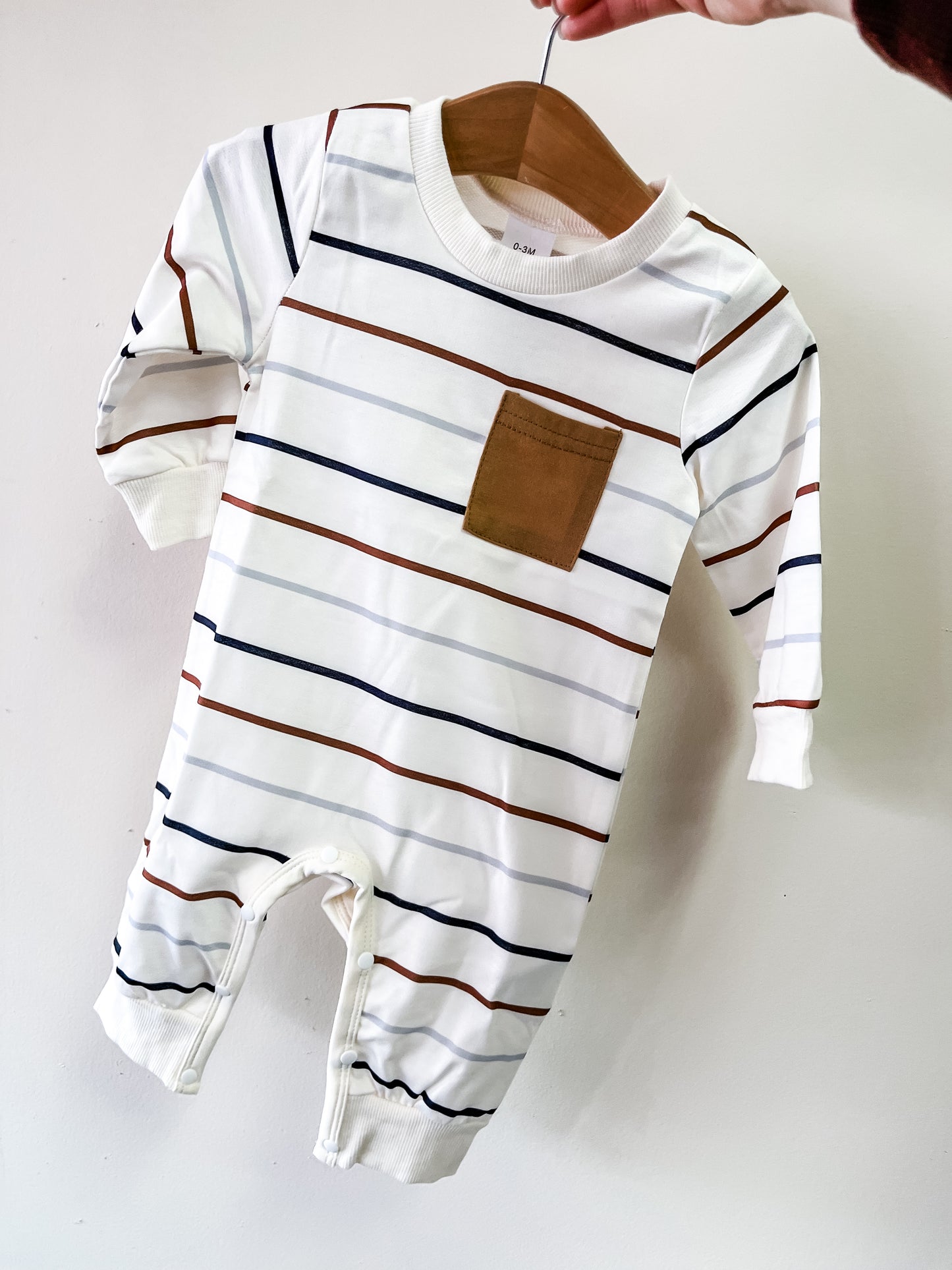 A hand displays Tiny Tot Bundles’ Neutral Stripe Pocket Romper, a white long-sleeve baby outfit with horizontal navy, brown, and orange stripes and a brown square chest pocket.