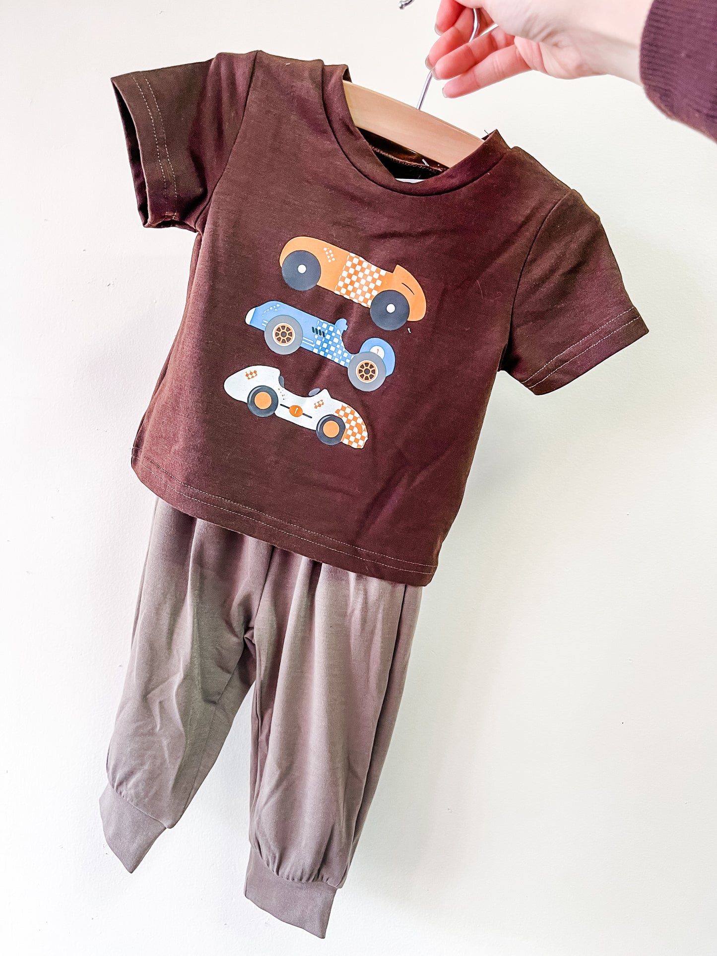 A hand displays the Tiny Tot Bundles Vintage Speedway Set: a brown short-sleeved toddler outfit with a shirt featuring three colorful race cars and coordinating lighter brown pants.