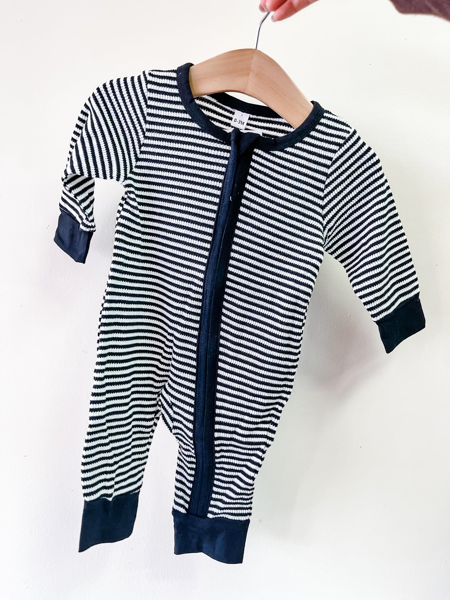 A hand holds the Tiny Tot Bundles Midnight Stripe Zip Romper, a baby-sized long-sleeve outfit with black and white stripes, solid dark cuffs and collar, and a front zipper.