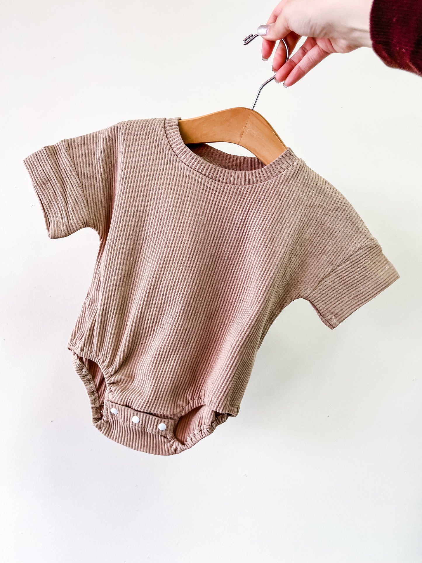A hand holds a wooden hanger with the Tiny Tot Bundles Brown Cozy Ribbed Bubble, a short-sleeve baby romper with snap buttons at the bottom, shown against a plain white background.