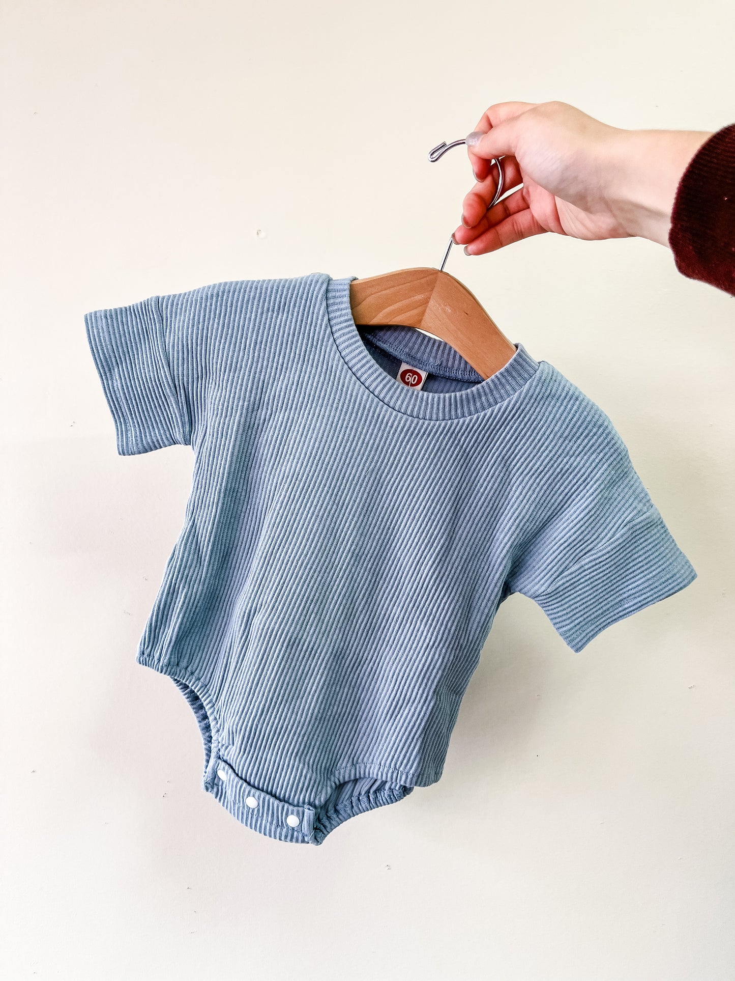 A hand holds a wooden hanger with the Tiny Tot Bundles Blue Cozy Ribbed Bubble, a short-sleeve, light blue baby onesie, against a plain light background.