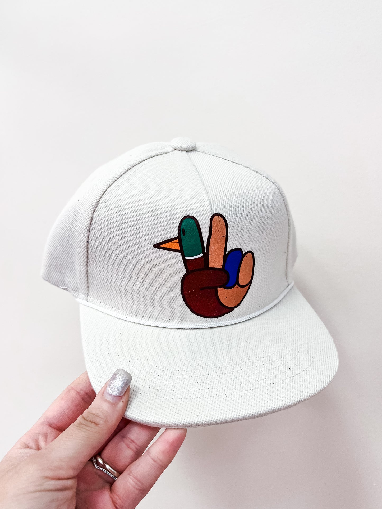 A hand holds the Lucky Duck Peace Hat by Thats Rad, a white cap with a colorful cartoon hand making a peace sign—one finger cleverly designed as a duck’s head.