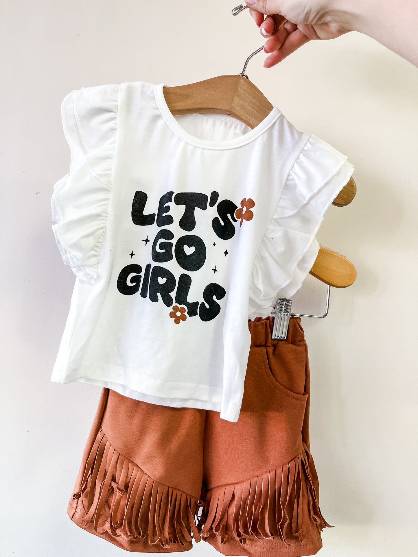 A hand holds the Let's Go Girls Ruffle Set by Tiny Tot Bundles: a kids outfit with a white ruffled-sleeve top featuring "LETS GO GIRLS" and orange flowers, paired with rust-colored fringe shorts.