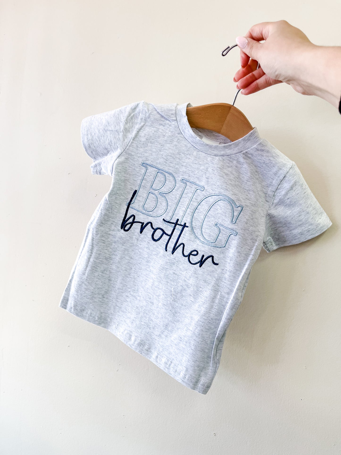 A hand holds up the Tiny Tot Bundles Big Brother Scripted Tee, a small gray toddler t-shirt with “BIG brother” in bold blue and black letters, displayed on a hanger against a plain light background.