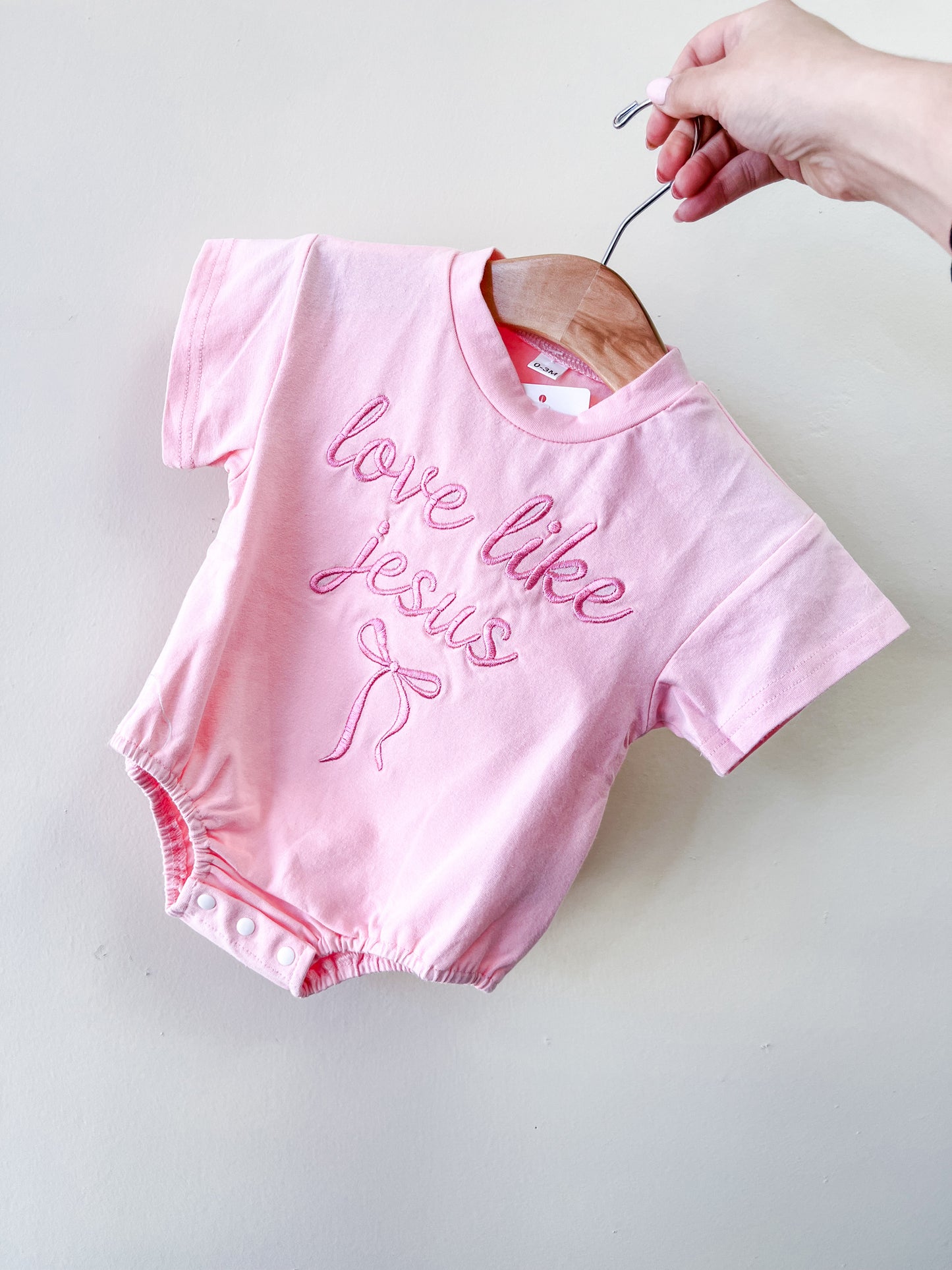 A hand holds a wooden hanger with the Tiny Tot Bundles Love Like Jesus Bubble, a pink baby romper featuring "love like Jesus" and a bow embroidered on the front, displayed against a plain light background.