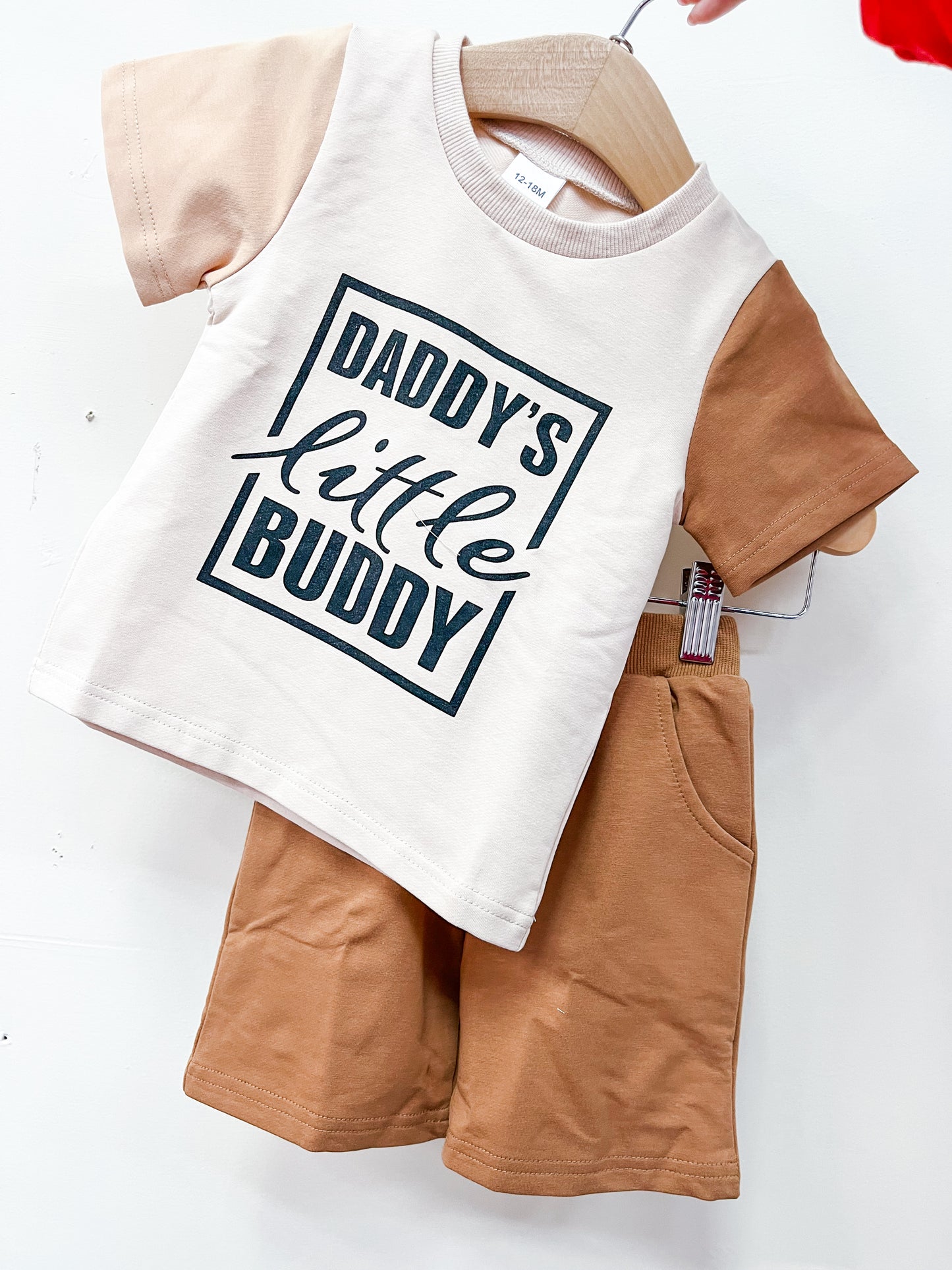 The Daddy's Little Buddy Short Set by Tiny Tot Bundles features a beige T-shirt with tan sleeves and "Daddy's little buddy" on the front, paired with matching tan shorts, displayed on a hanger.