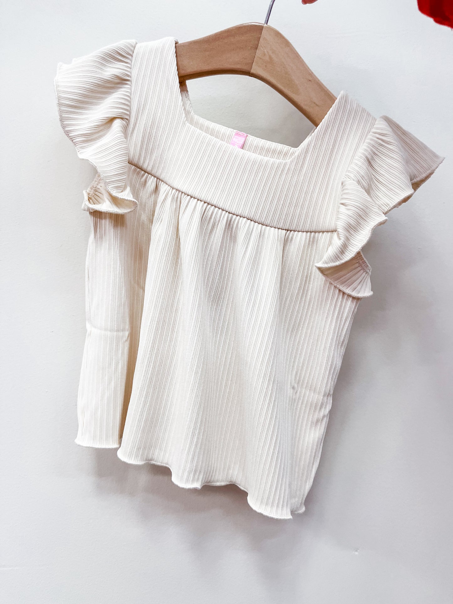 The Buttercream Breeze Blouse by Cutie Patootie is a cream-colored, short-sleeve top with subtle vertical stripes, ruffled sleeves, and a square neckline, displayed on a wooden hanger against a white background.