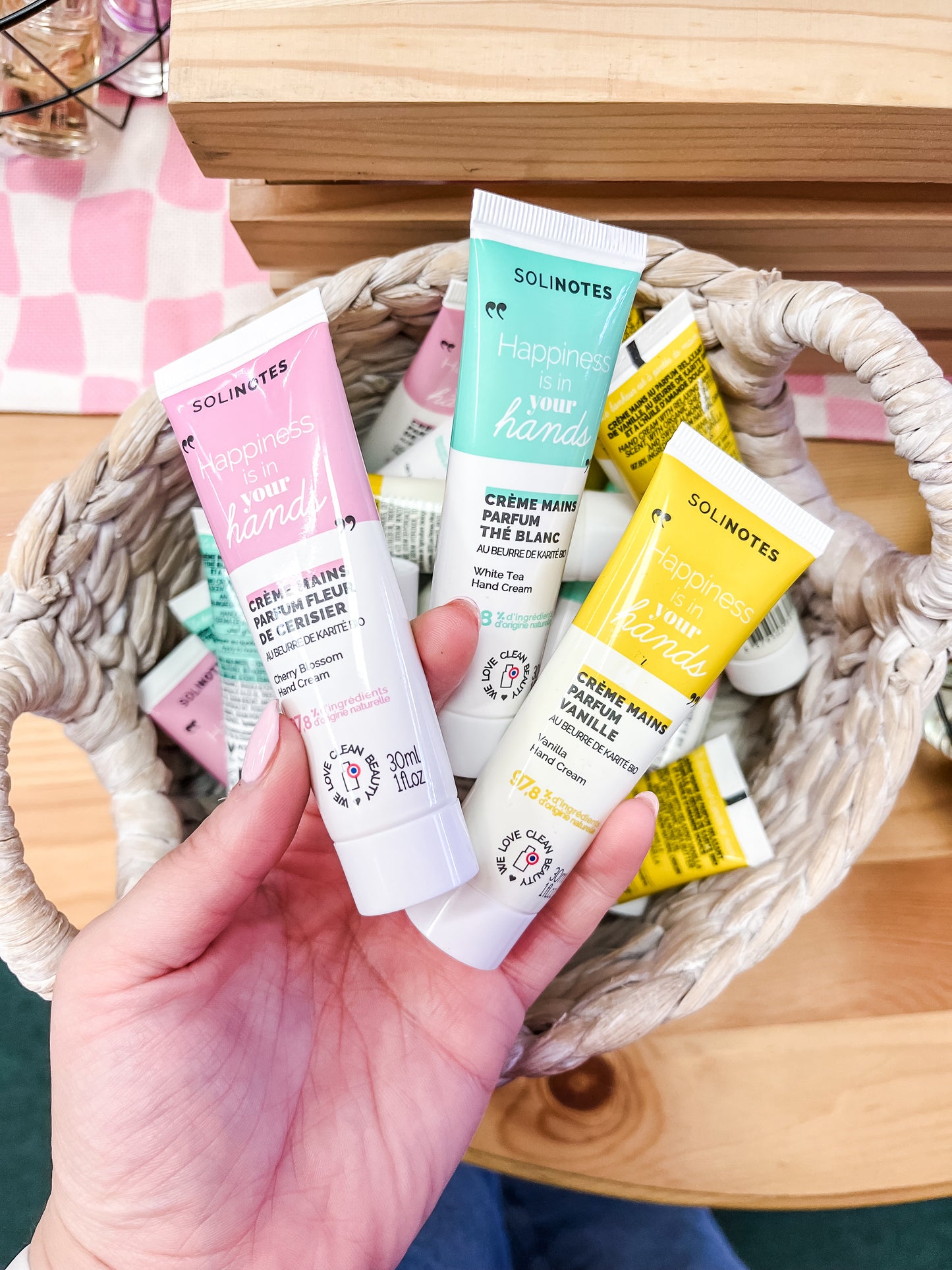 A hand holds two Solinotes Hand Creme tubes above a basket filled with more pastel-colored tubes from Solinotes, all displayed on a wooden surface.