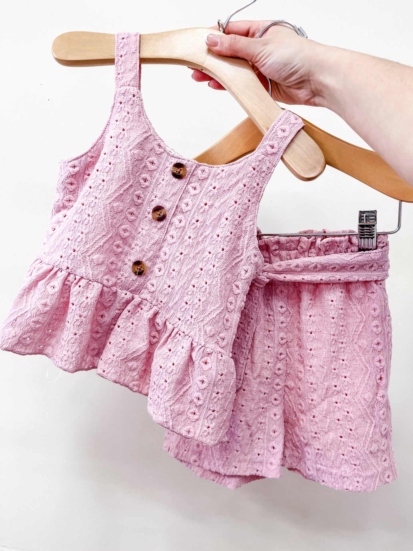 A hand holds a hanger with the Tiny Tot Bundles Dusty Rose Set, featuring a pink, eyelet-patterned sleeveless toddler top with wooden buttons and a ruffled hem, next to matching shorts on another hanger.