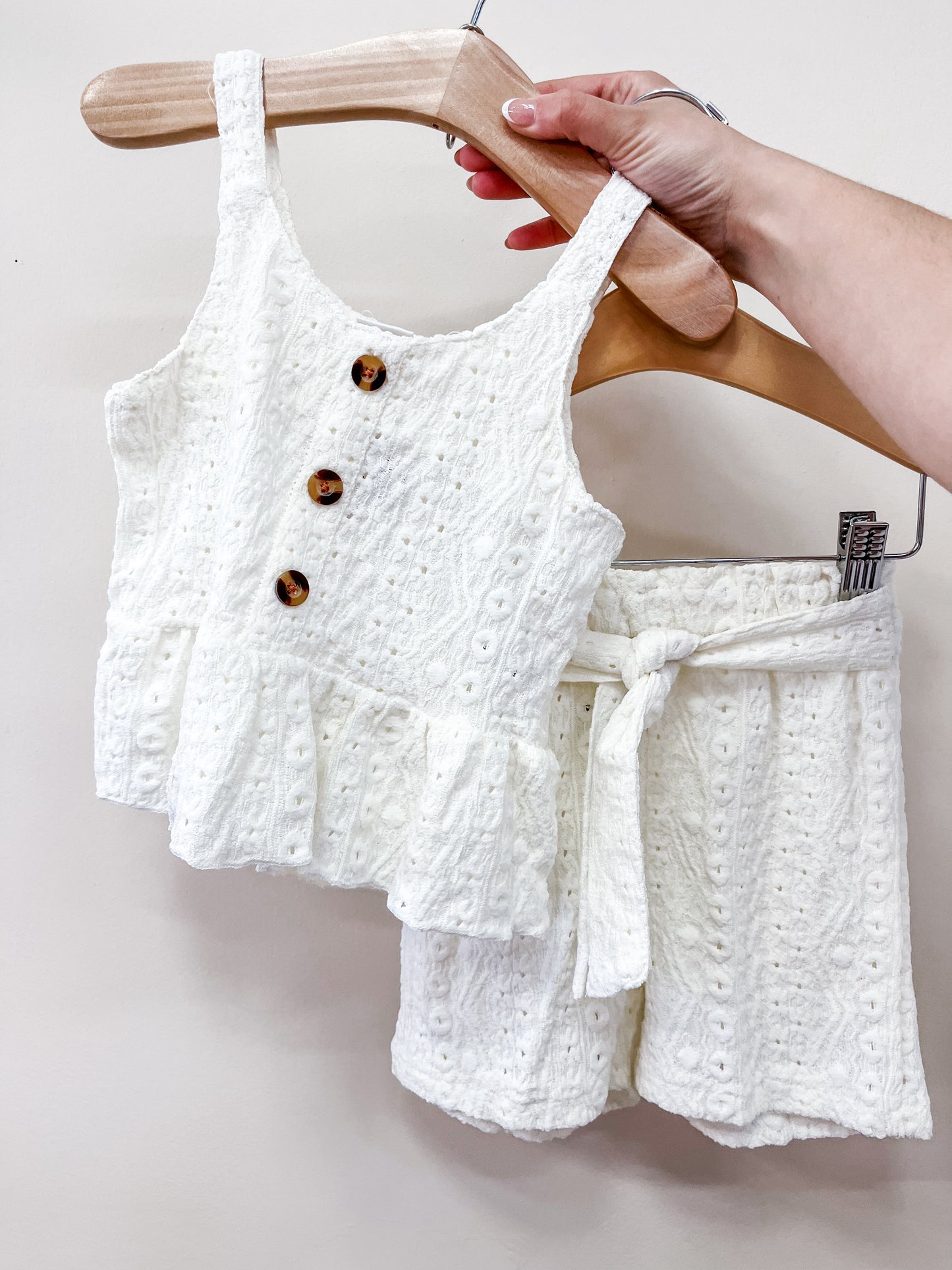 A hand holds a Tiny Tot Bundles Sweet Cream Tank Set: a sleeveless ruffled white top with three brown buttons and matching shorts with a tie belt, displayed on a wooden hanger against a light background.
