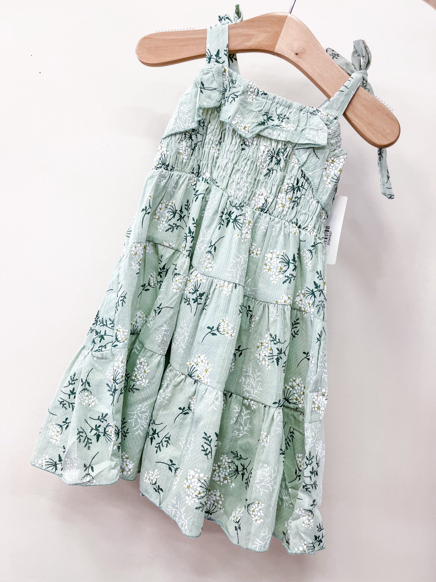 The Tiny Tot Bundles Sage Mellow Dress is a young girl's light green, sleeveless sundress with floral print, ruffled top edge, tiered design, and adjustable shoulder ties, shown hanging on a wooden hanger.