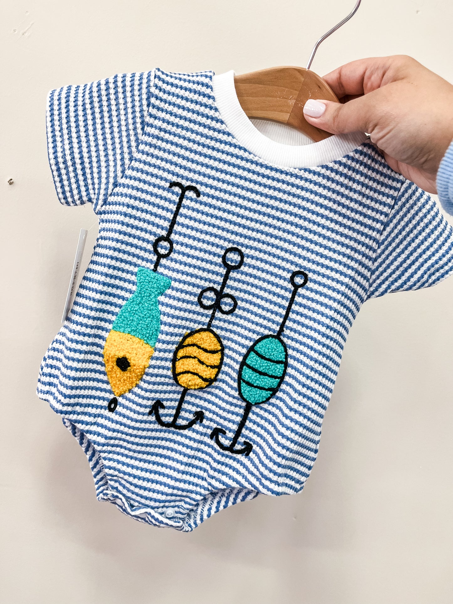 A hand holds the Catch Of The Day Bubble by Tiny Tot Bundles, a blue and white striped baby onesie with short sleeves, featuring colorful embroidered fishing lures on the front.