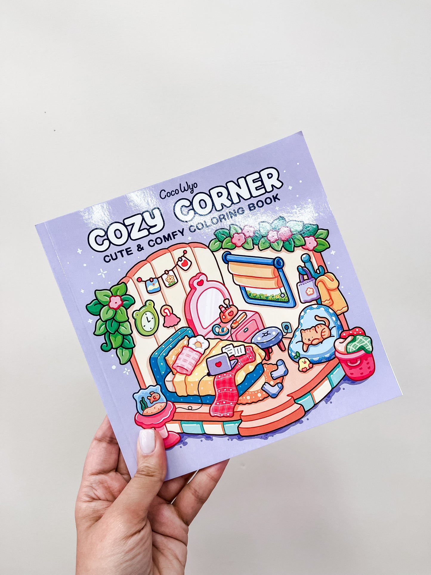 A hand holds a pastel-colored Cozy Corner Coloring Book by Tiny Tot Bundles, featuring an illustrated cozy room with plants, books, a cat, tea, and knitting supplies on the cover.