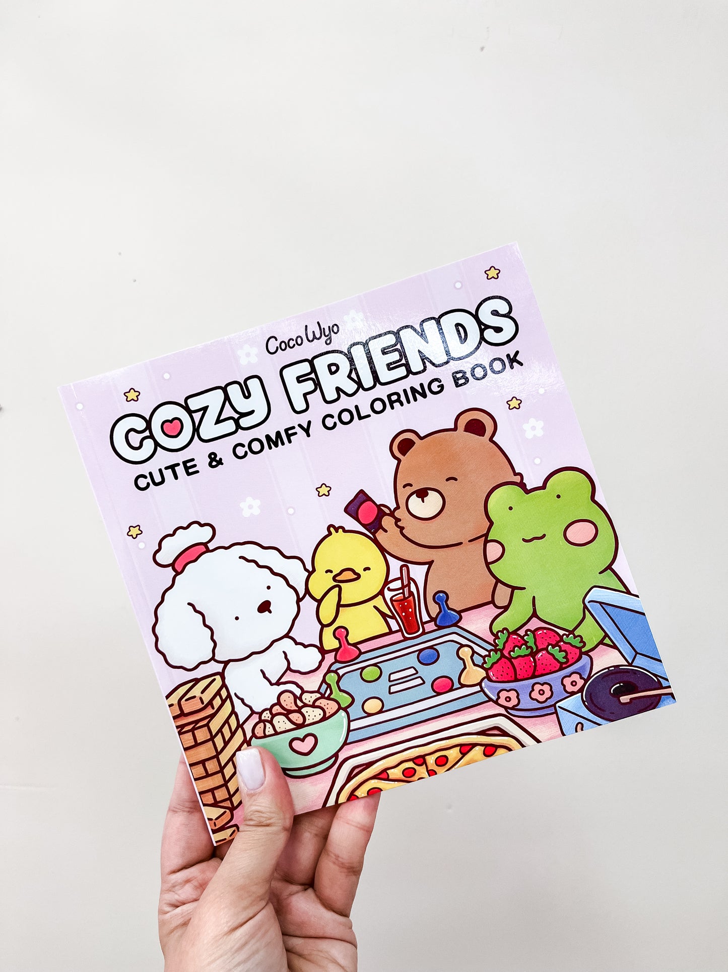 A hand holds a Tiny Tot Bundles Cozy Friends Coloring Book featuring cartoon animals like a dog, duck, bear, and frog sitting around a table with food and art supplies on a pastel background.
