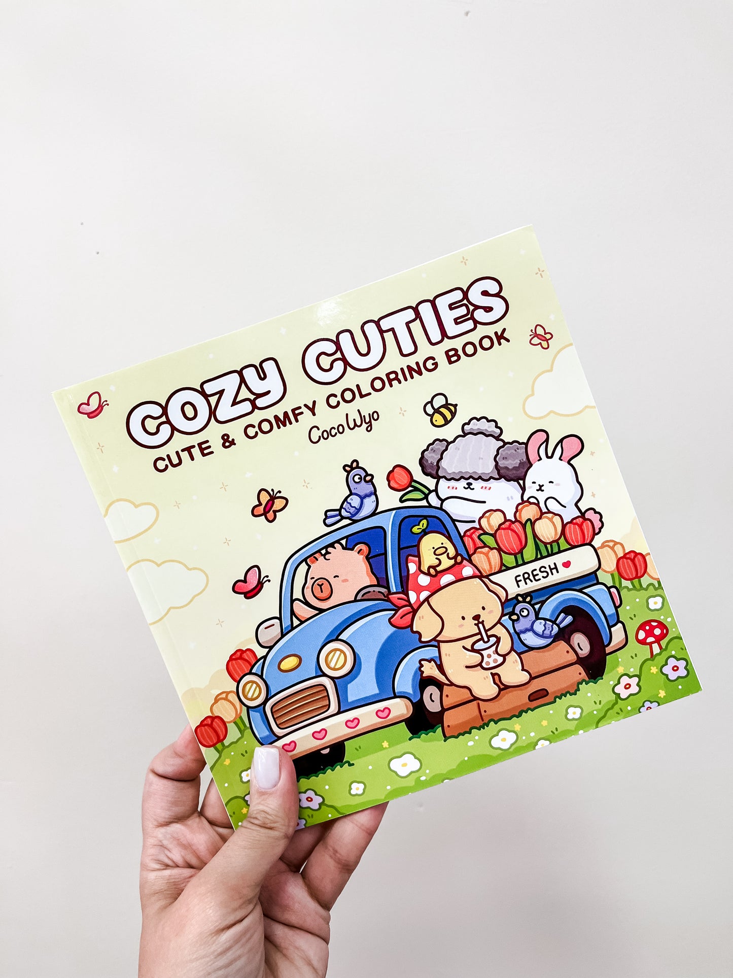 A hand holds the Cozy Cuties Coloring Book by Tiny Tot Bundles, featuring cartoon animals with apples around a blue car in a grassy outdoor scene on the cover.