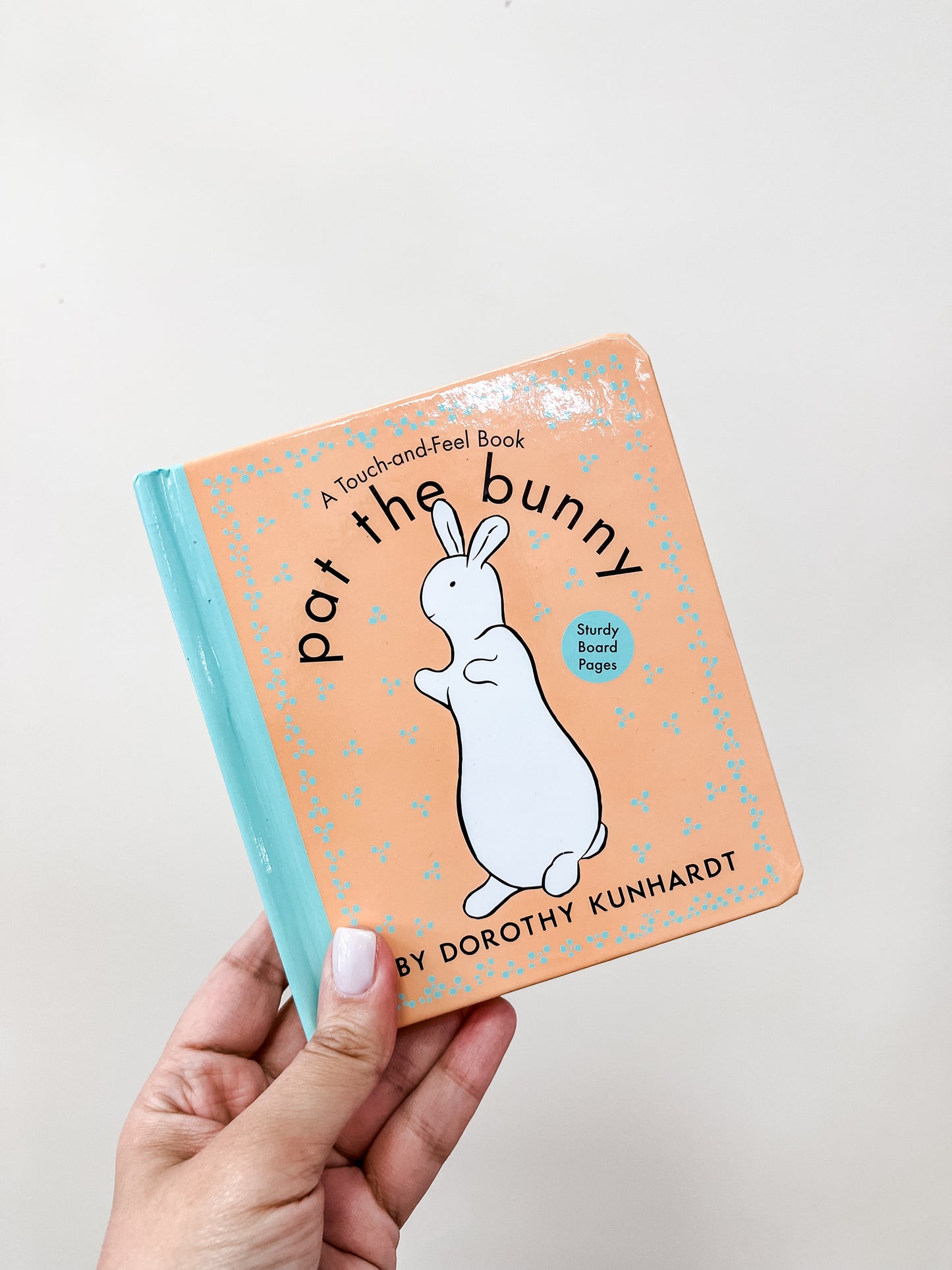 A hand holds the Pat the Bunny Book by Tiny Tot Bundles. The peach cover with a blue spine shows a white bunny and reads "A Touch-and-Feel Book" and "Sturdy Board Pages.