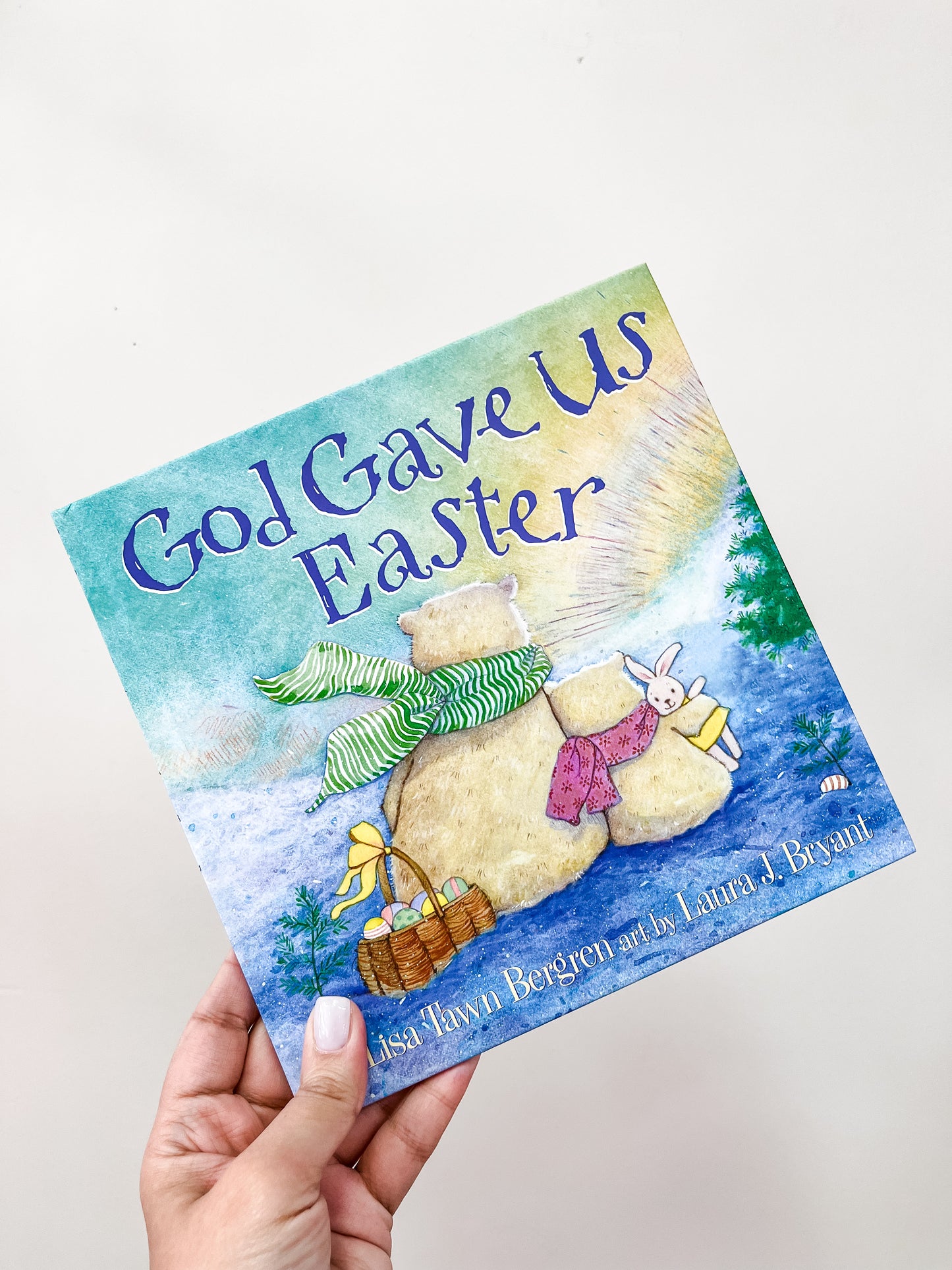 A hand holds the God Gave Us Easter Book by Tiny Tot Bundles, featuring an illustrated bear family, basket, and toy rabbit on its snowy cover.