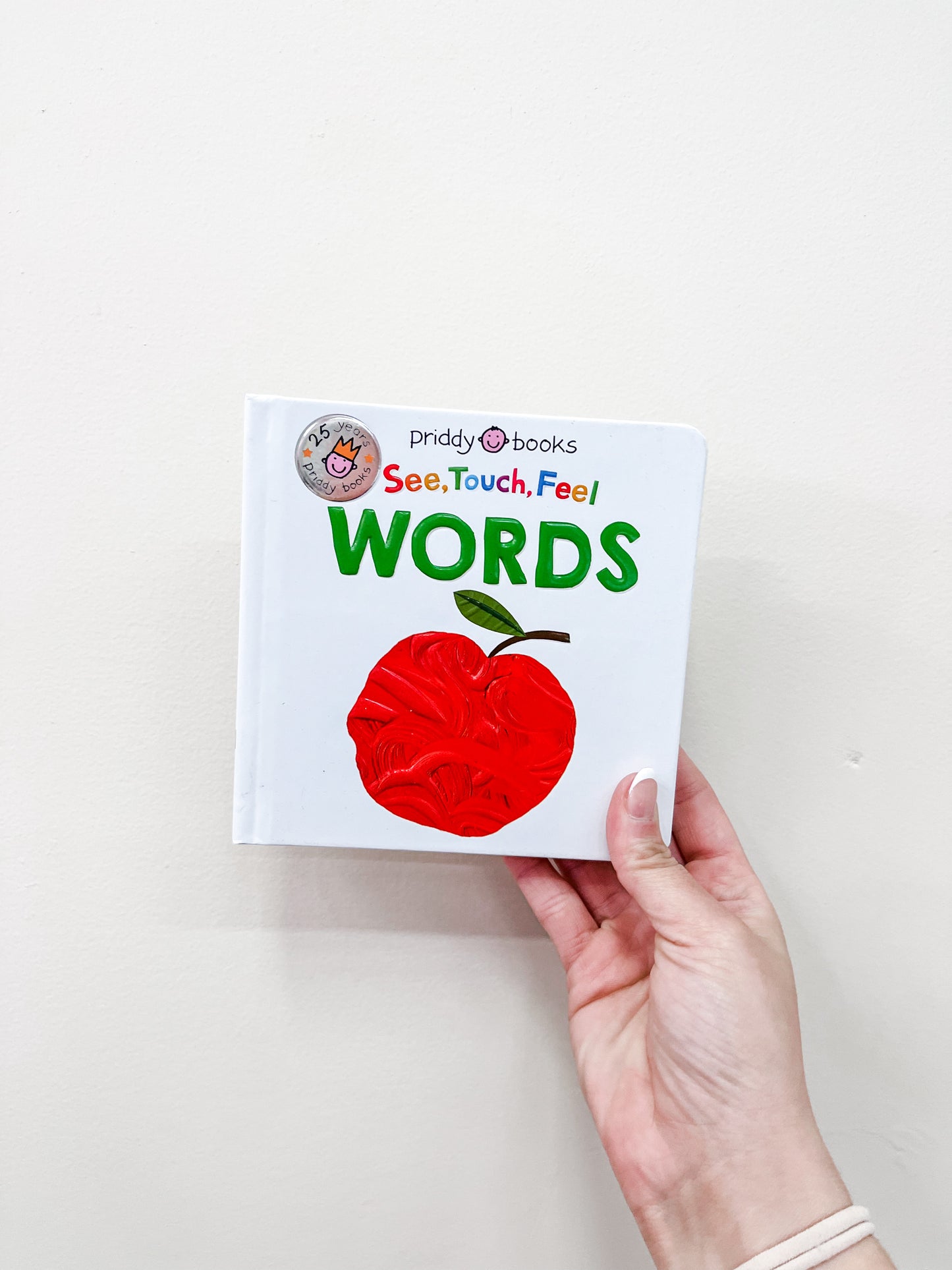 A hand holds a children's board book titled See Touch Feel - Words Book by Tiny Tot Bundles. The cover shows a large red apple with textured details on a white background.