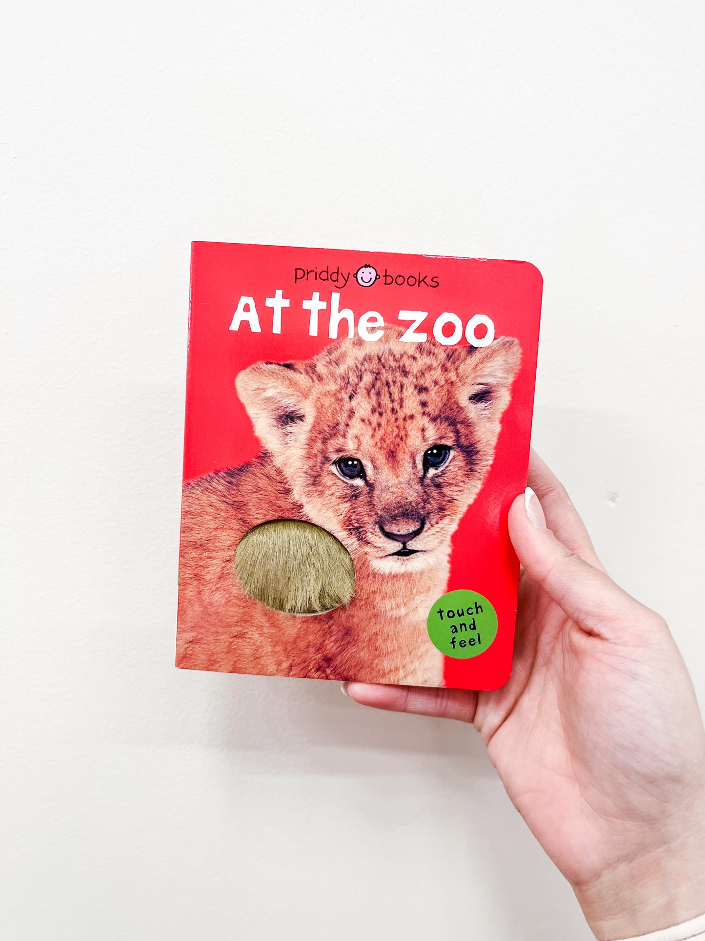 A hand holds the Macmillan Publishers "T & F - At the Zoo Book," a red children's book with a baby lion and furry touch-and-feel patch on the cover.
