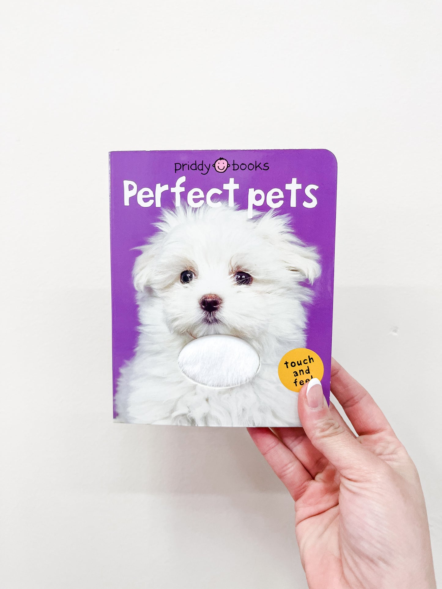 A hand holds the Macmillan Publishers T & F - Perfect Pets Book, featuring a fluffy white puppy on its purple cover and a textured patch for touching.